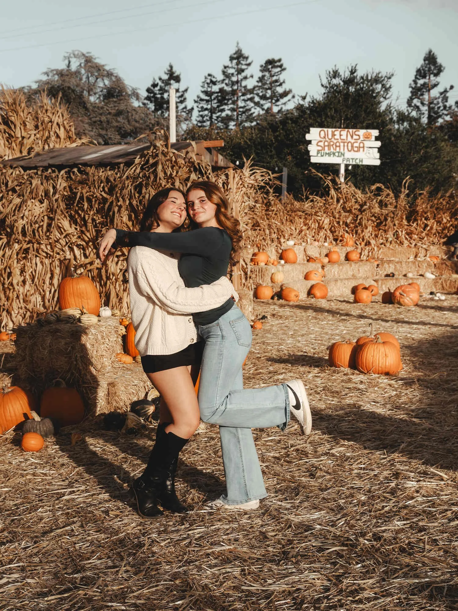 Two young women hugging and smiling at a pumpkin patch with pumpkins around them, hay bales, and a sign that reads 'Queens Saratoga Pumpkin Patch'.