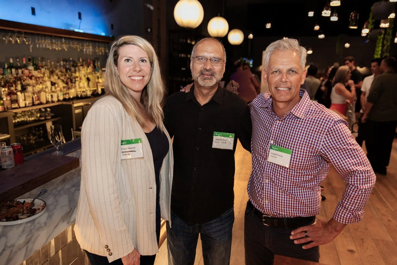 Three people at a social event, standing in a bar or restaurant with a well-stocked bar behind them, smiling at the camera.