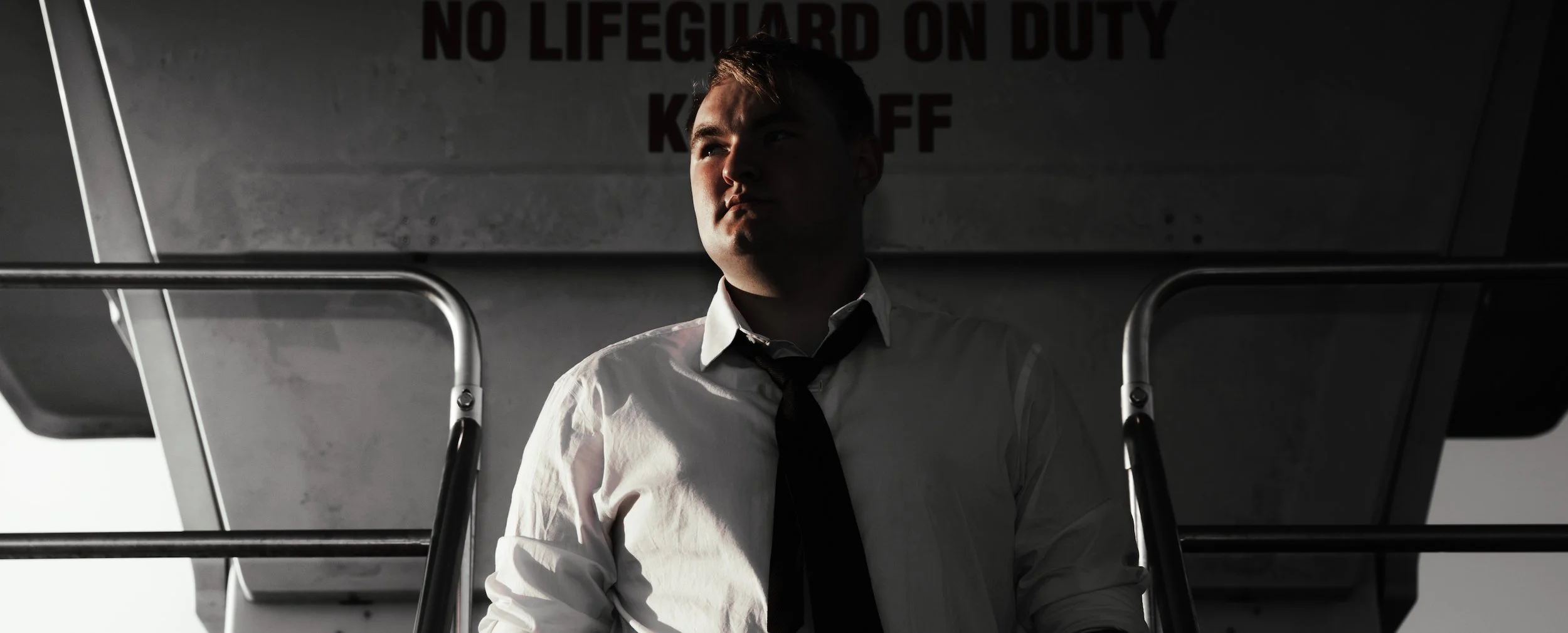 A young man in a white shirt and black tie looking thoughtful, standing under an overpass with a 'No Lifeguard on Duty' sign behind him.