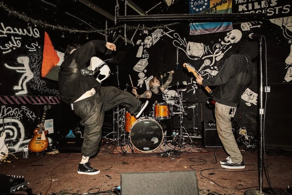 Three band members performing live on stage with graffiti background, drums, and musical instruments.