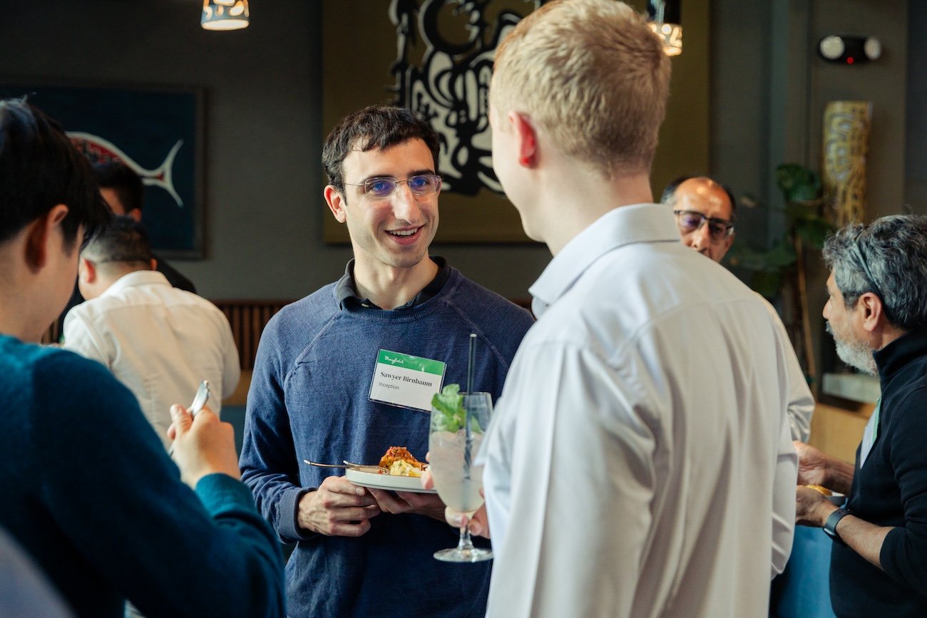 A man with glasses and a name tag talking with a blonde man holding a plate and drink at a social event or conference.