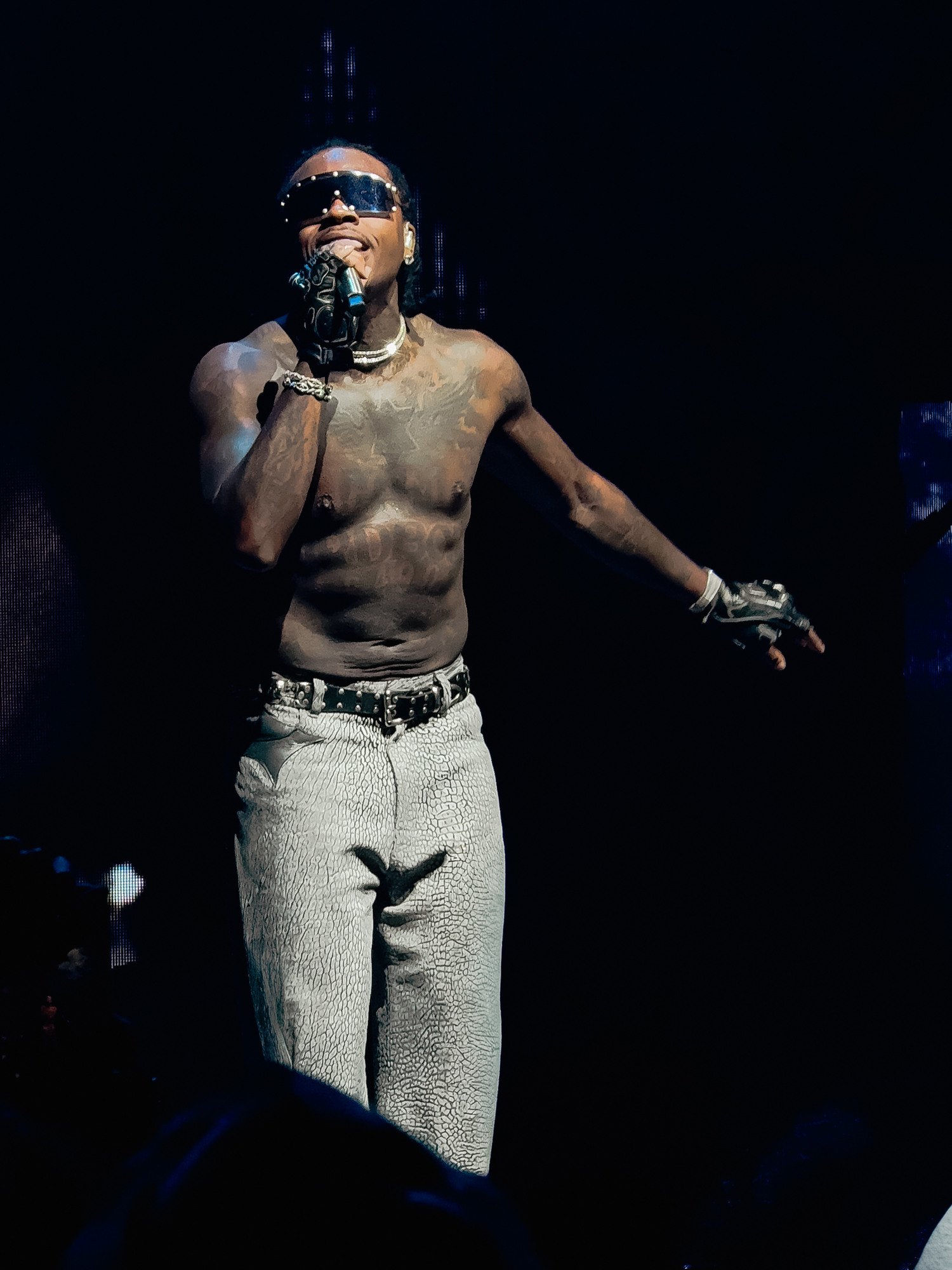 A shirtless male performer wearing sunglasses, earrings, and jewelry, holding a microphone, on a dark stage with minimal background lighting.