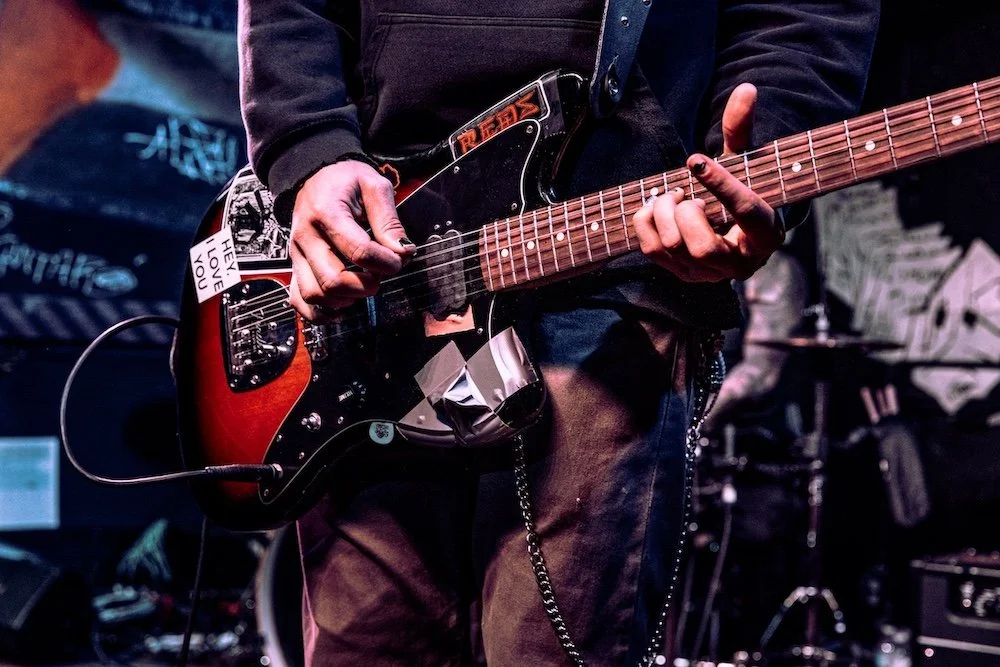Close-up of a person playing an electric guitar on stage, with graffiti-style artwork in the background.
