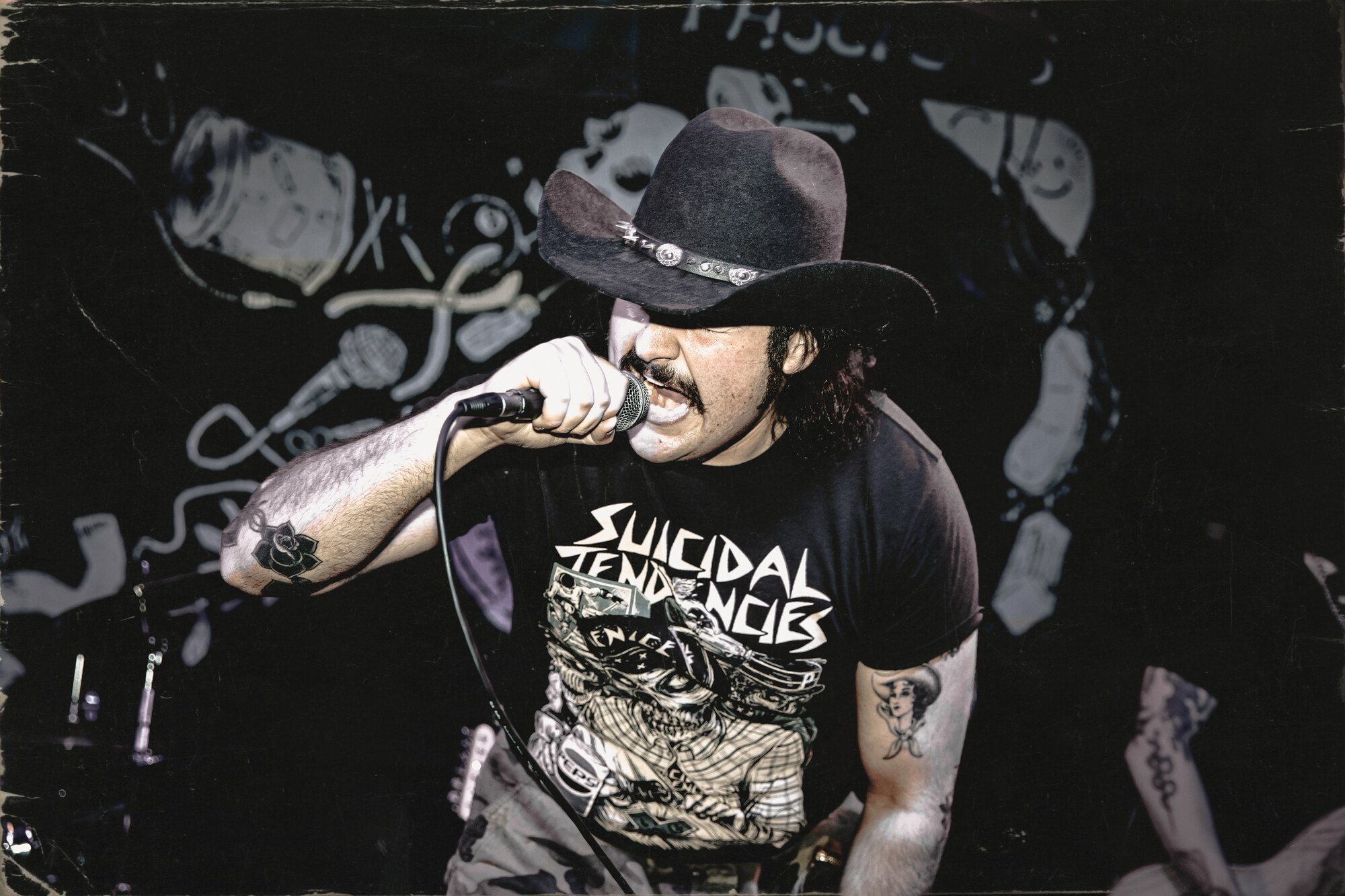 A male singer wearing a large black cowboy hat, a black T-shirt with the words "Suicidal Tendencies," singing into a microphone on stage, with tattoos on his arm, including a rose and a woman, against a dark background with graffiti-style artwork.