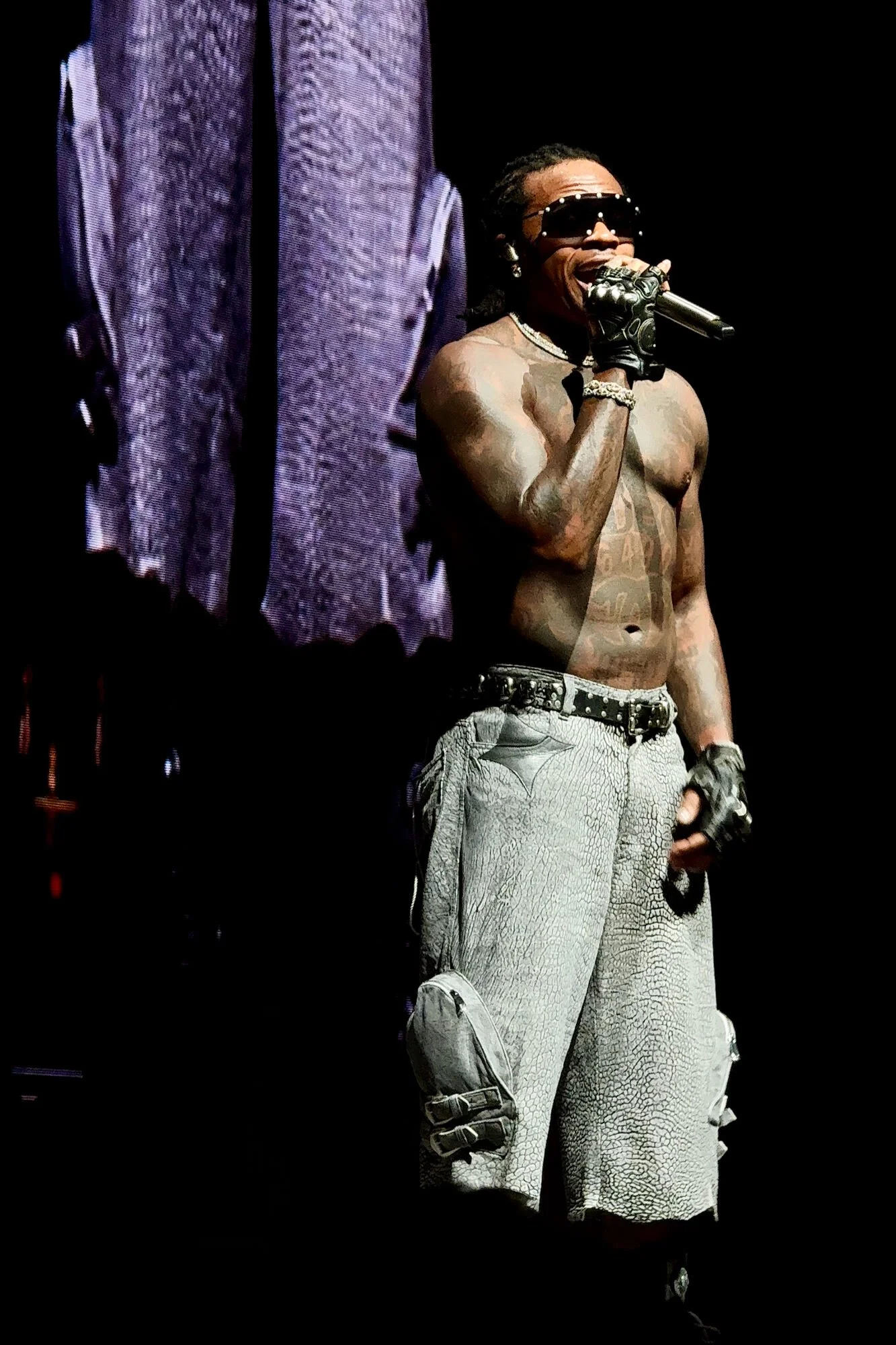 A shirtless man with tattoos, wearing sunglasses, gloves, and cargo shorts, singing into a microphone on stage.