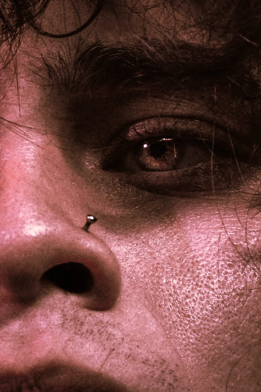 Close-up of a person's face, showing one eye with a brown iris, a nose piercing, and skin with visible pores and facial hair, under dim lighting.