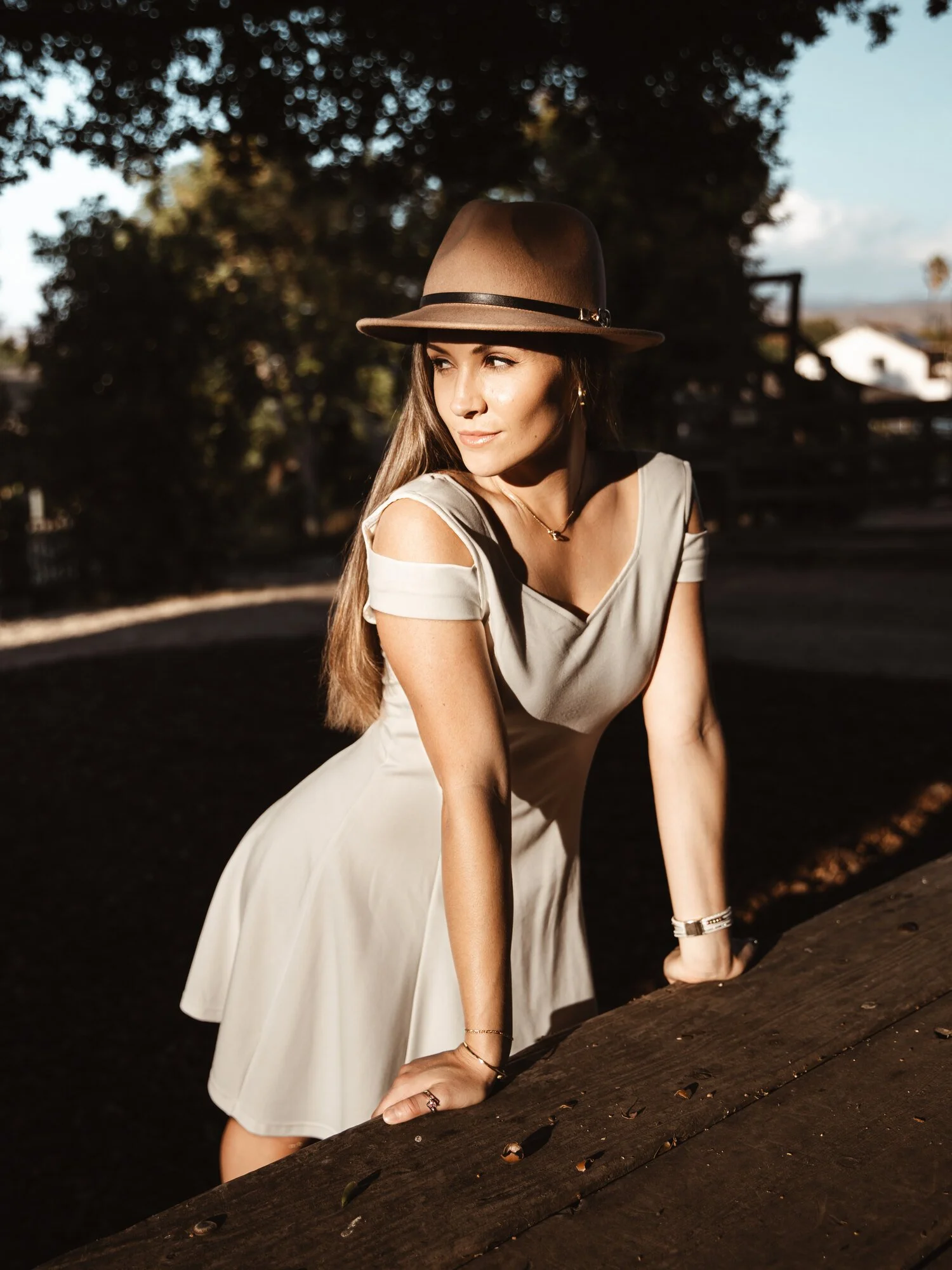 A young woman with long brown hair wearing a beige dress with shoulder cutouts, a brown fedora hat, and jewelry, leaning on a wooden surface outdoors during the daytime with trees and blue sky in the background.