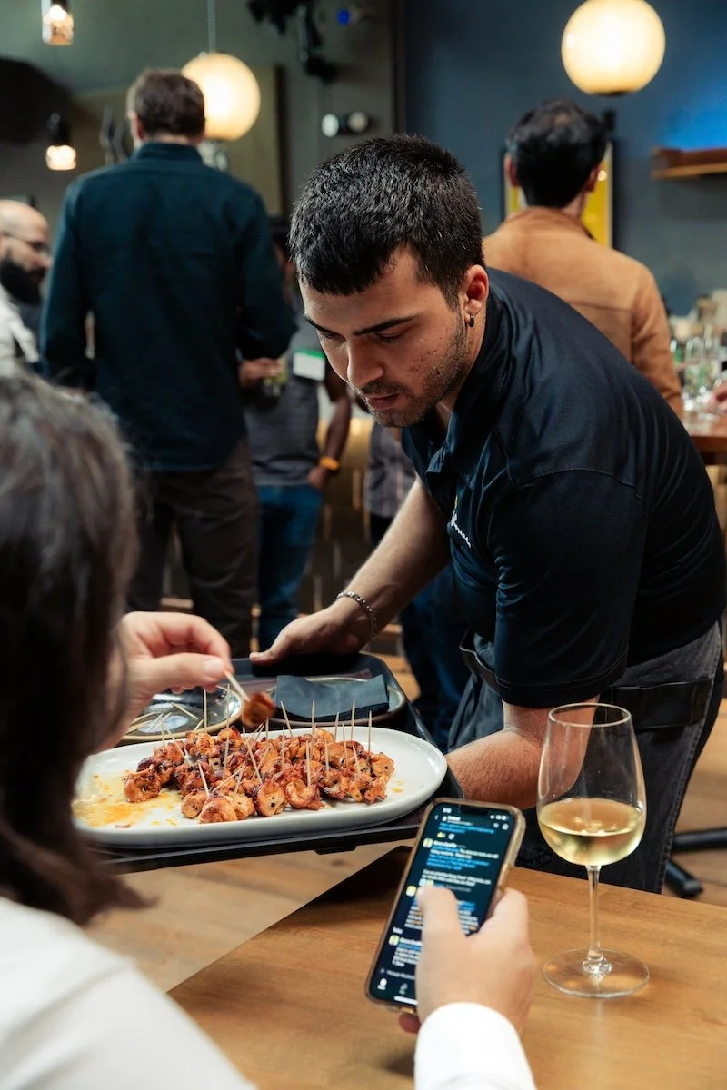 A server delivers a tray of nachos to a customer in a restaurant, with a glass of white wine and a person holding a phone in the foreground.