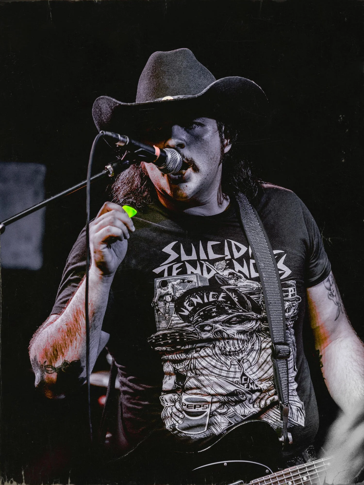 Musician wearing a cowboy hat and black T-shirt singing into a microphone on stage.