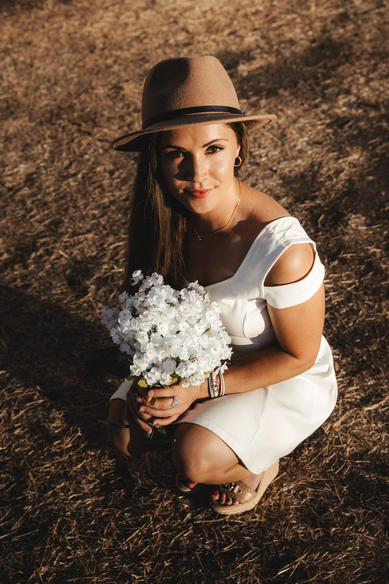 A woman in a white dress and tan hat kneeling on dry ground, holding a bouquet of white flowers, with sunlight casting shadows on her face.