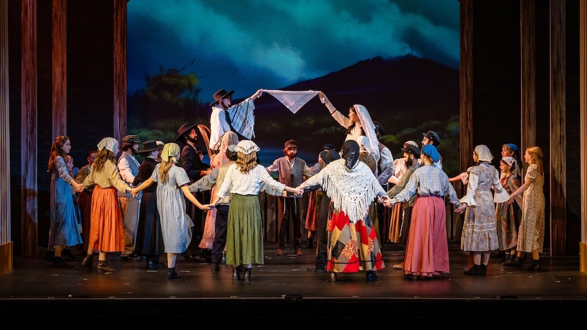 A theatrical play with multiple actors on stage dressed in rustic, historical costumes, performing in a circle with two actors elevated in the center holding a cloth or shawl, set against a painted landscape background.