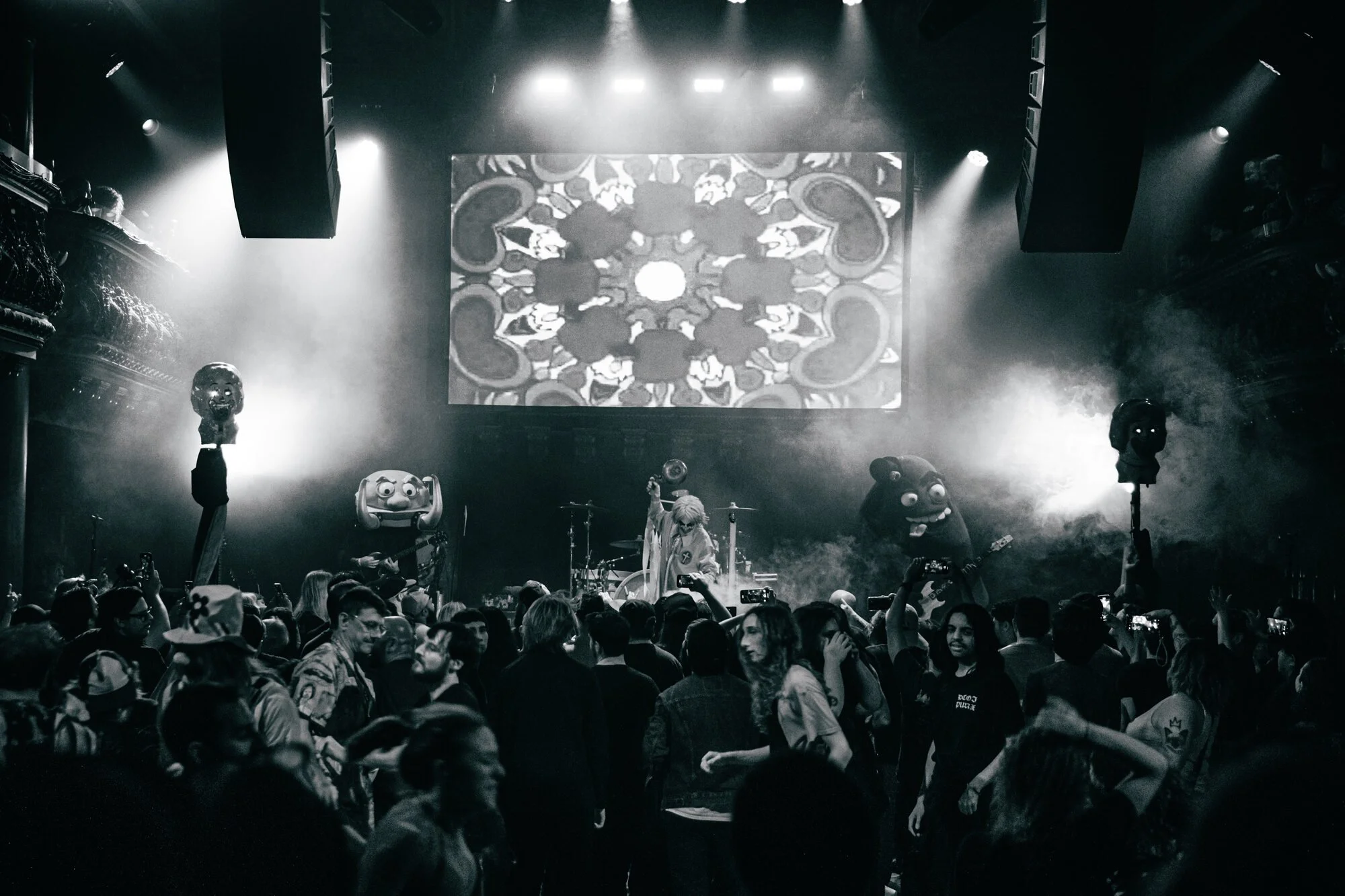 Black and white photo of a concert stage with a band performing, audience dancing, and giant puppets or mascot figures on either side of the stage.