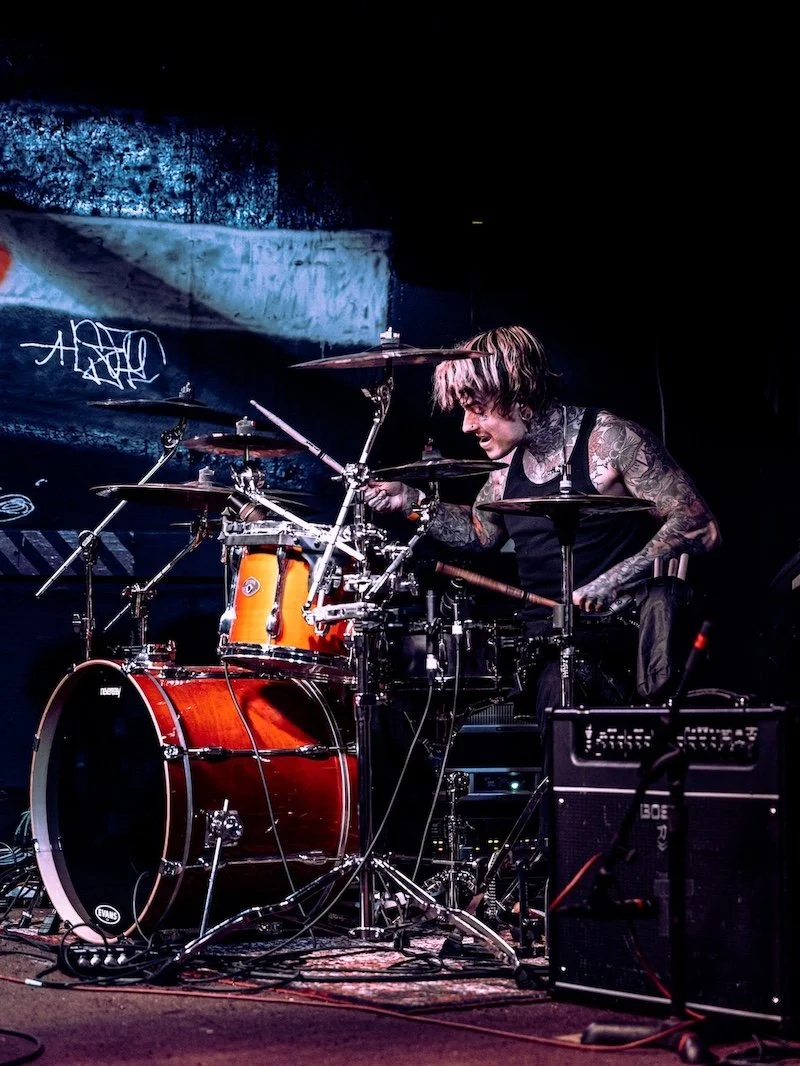A tattooed drummer playing an orange drum kit on stage with a dark background.