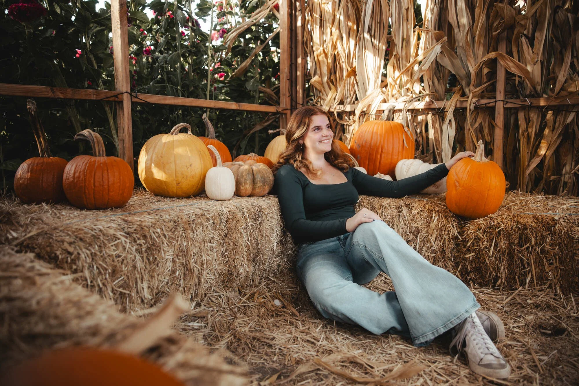 A young woman sits on straw beside pumpkins of various colors and sizes at a pumpkin patch, with dried corn stalks in the background, smiling and looking cheerful.