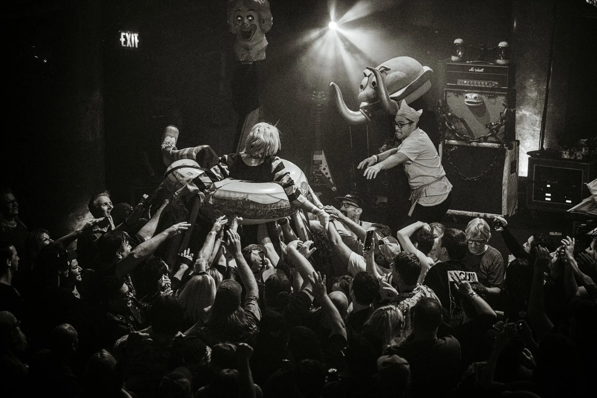 Crowd of people at a concert or party, reaching out towards individuals on stage. One person is in a rubber ring, and others are dressed in novelty costumes, including a person with a paper crown. The scene is lively with raised hands and smiling fac