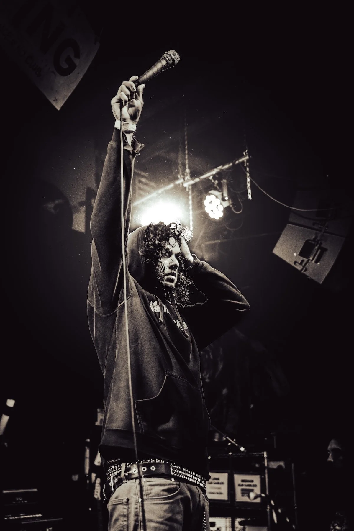A black-and-white photo of a young man with curly hair holding a microphone up in the air, on stage during a concert.