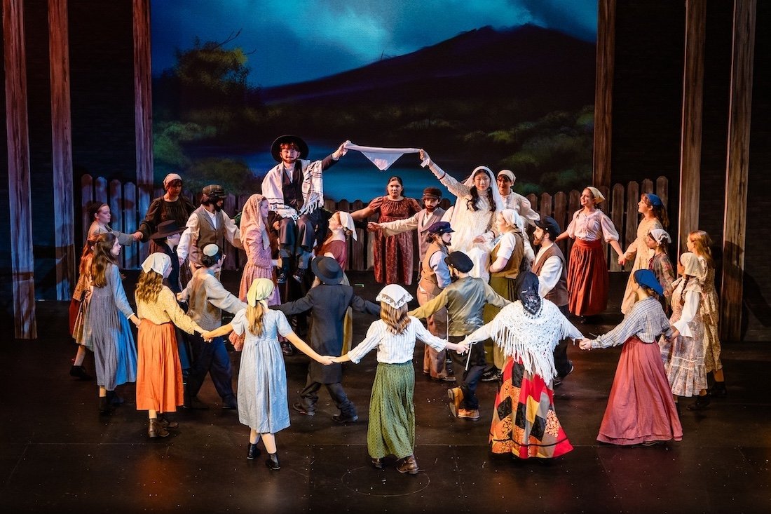 Theater cast performing a traditional folk dance on stage, with a painted outdoor scene backdrop