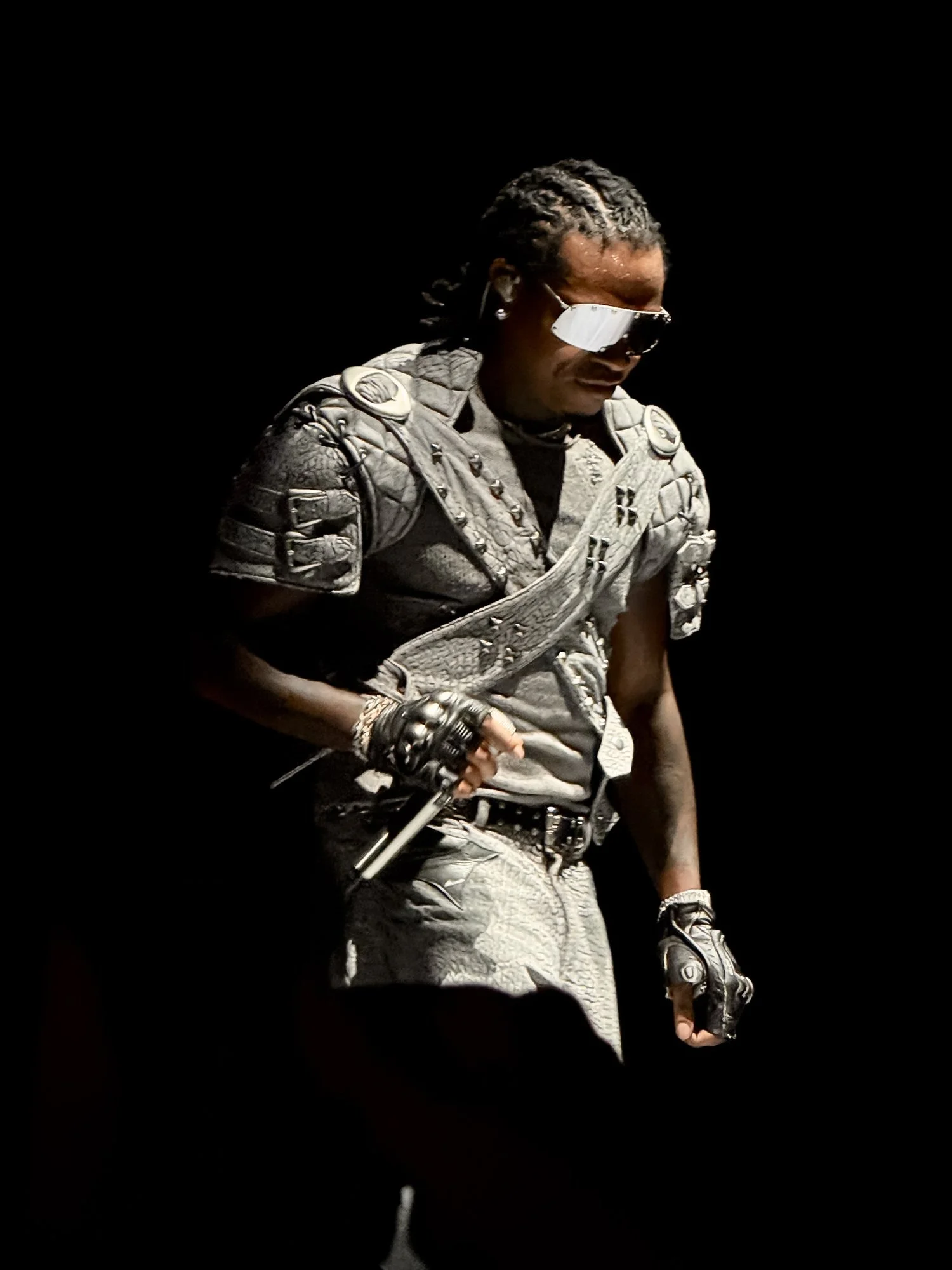 Person dressed in futuristic or warrior-style costume, wearing sunglasses, holding a prop gun, with a black background.