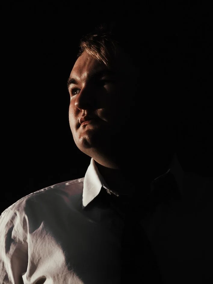 A man wearing a white shirt and black tie, illuminated from the left side, with the rest of his face and body in shadow, against a dark background.