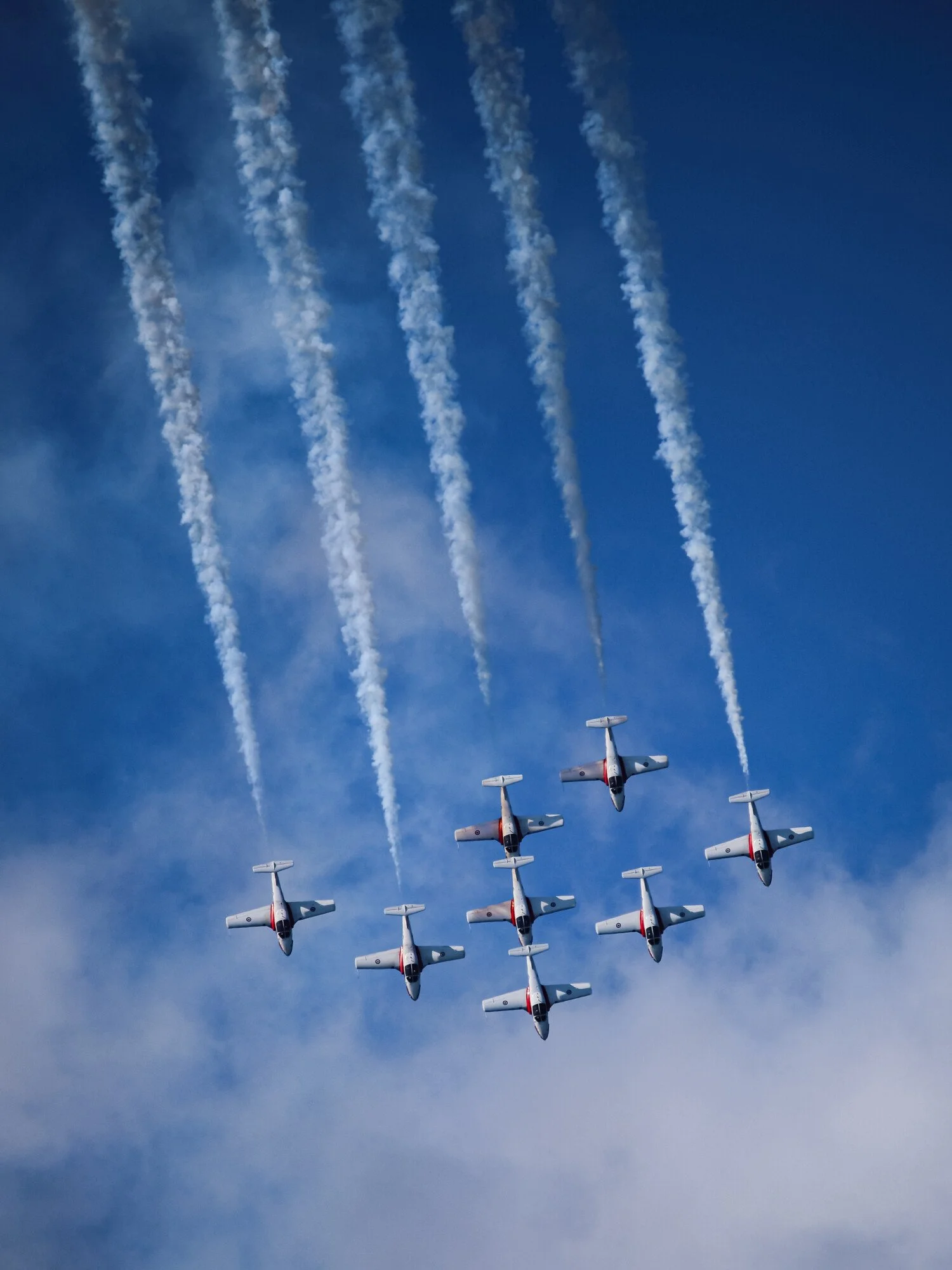 Seven fighter jets flying in a tight formation releasing white smoke trails against a blue sky with wispy clouds.