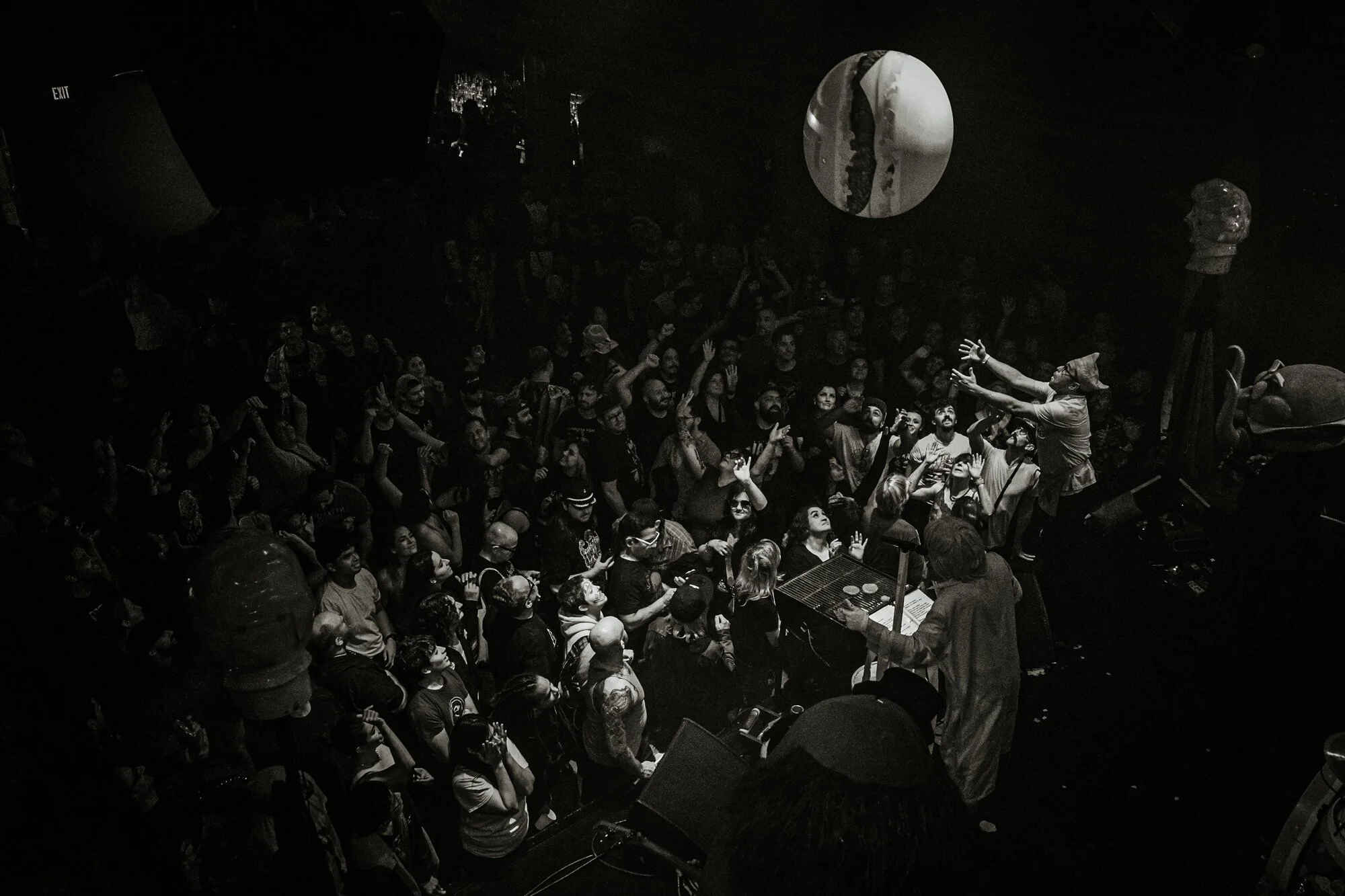 Black and white photo of a crowded concert with a performer engaging with the audience on stage, surrounded by various decorations including a large human head sculpture and a globe.