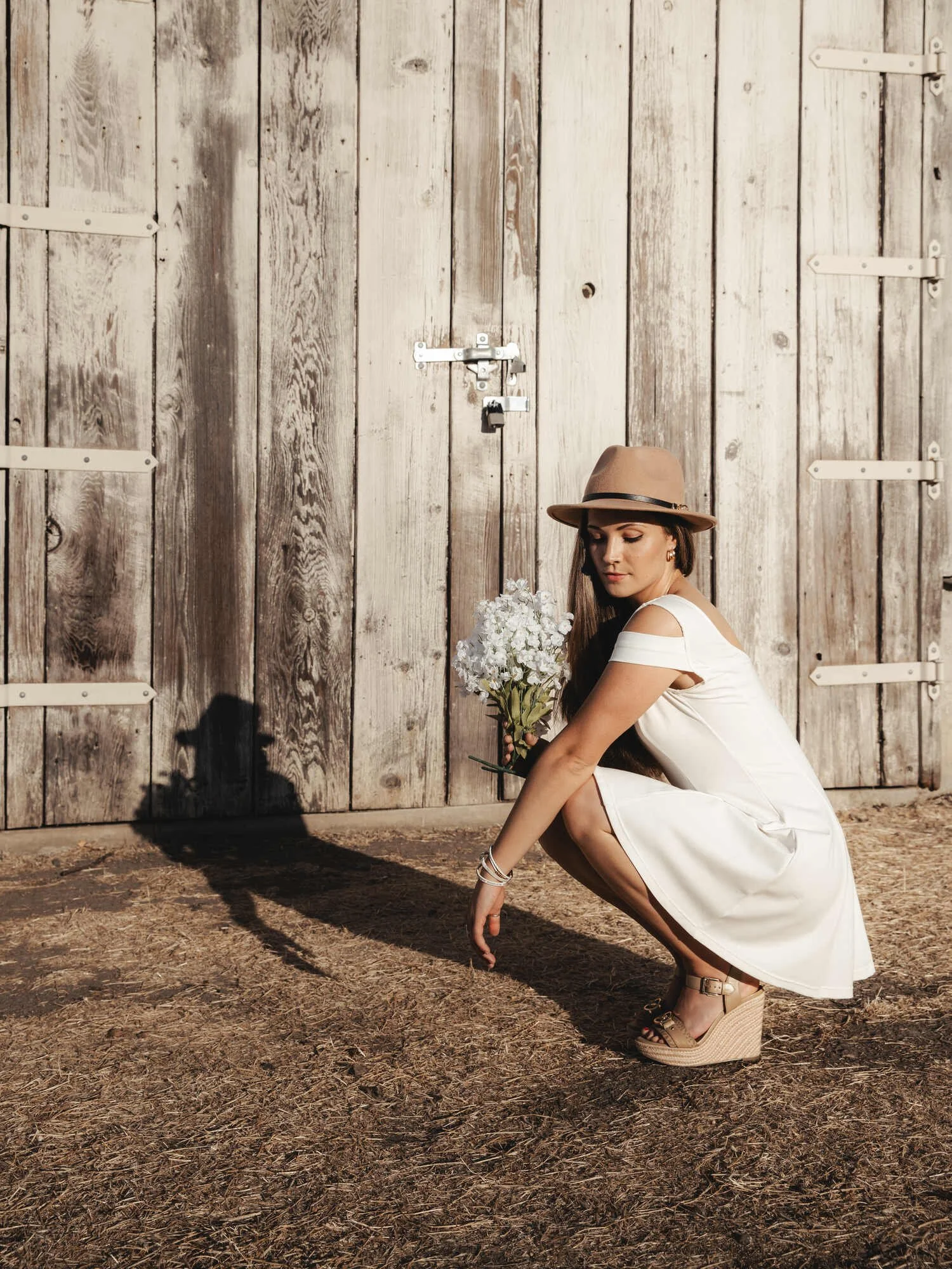 A woman in a white dress and tan wedge sandals squats on the dirt ground in front of a weathered wooden barn door, holding a bouquet of white flowers, wearing a tan fedora hat, with her shadow cast on the door.