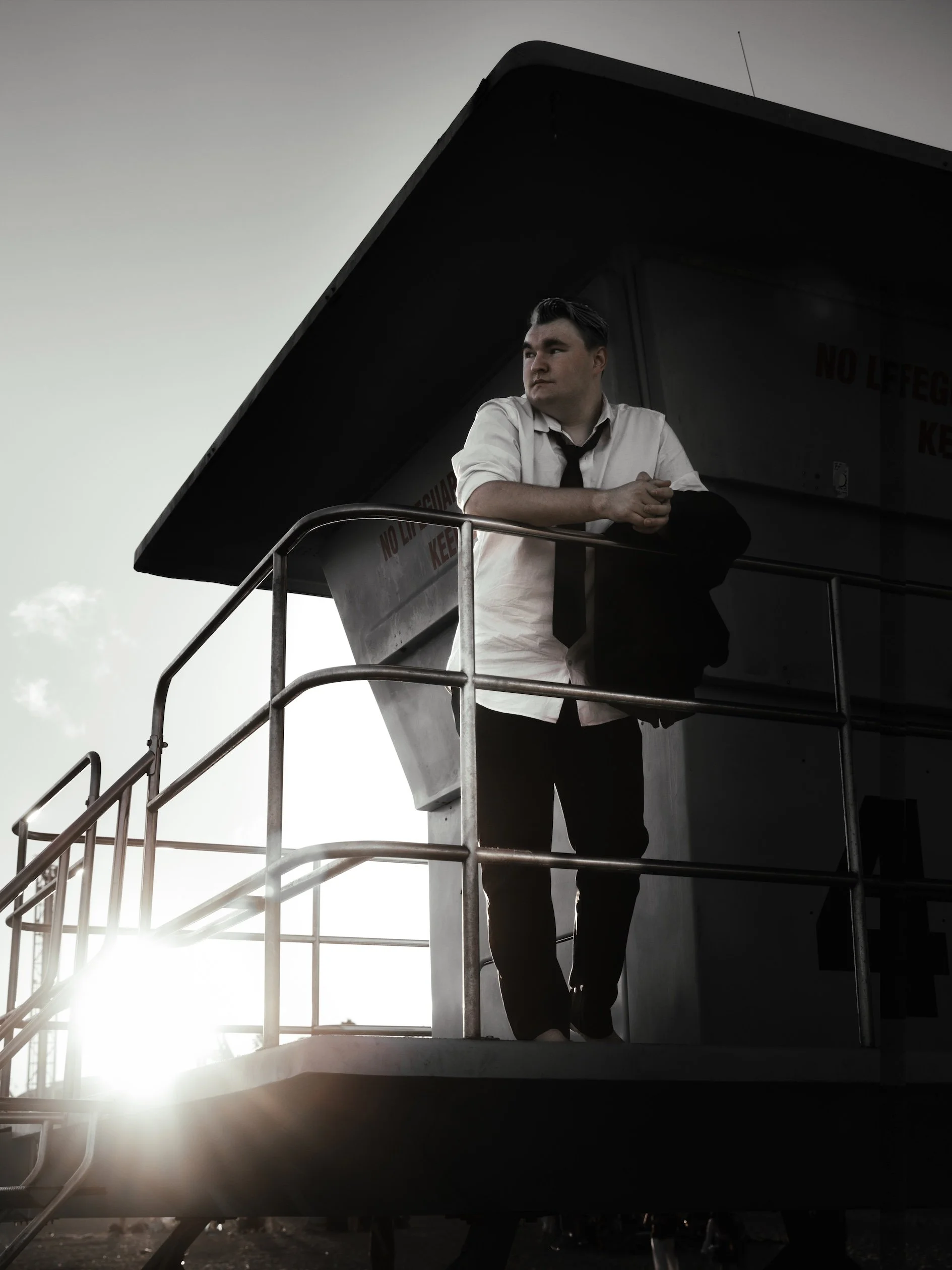 A man in a white shirt and black tie standing on a boat's observation deck with a railing, looking away into the distance, with the sun setting behind him.