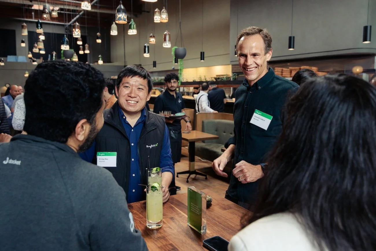 Four people having a conversation at a restaurant or bar, with two men standing and two people with backs to the camera seated at the table. The man on the left is holding a drink, and the atmosphere is lively and modern.