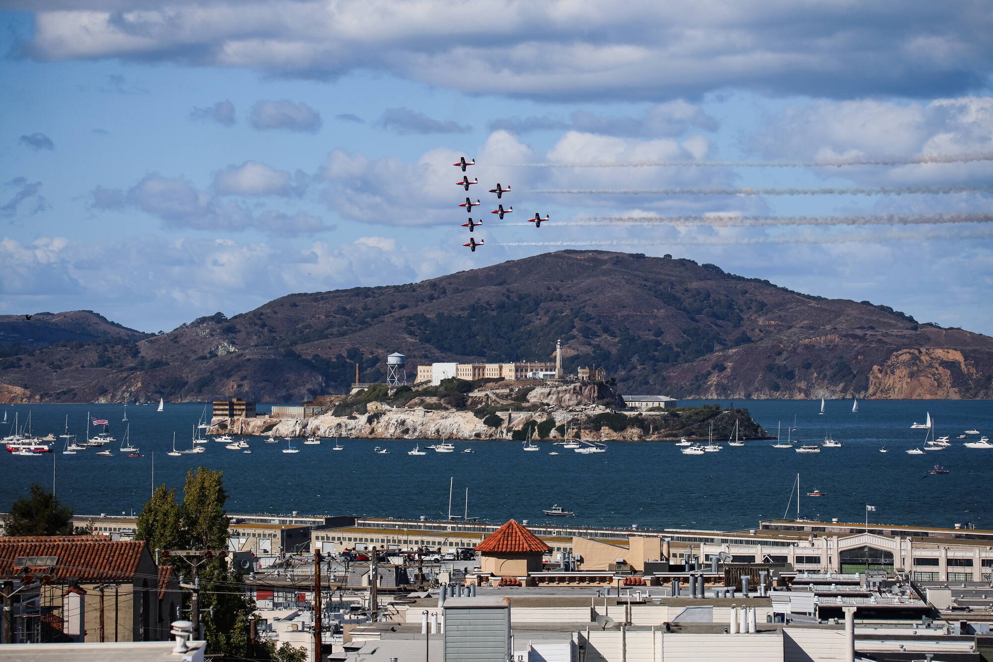 Fighter jets flying in formation over Alcatraz Island, with sailboats in the bay and city rooftops in the foreground.