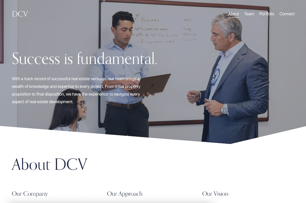 Business professionals in a meeting room, with a man in a gray suit speaking and a woman with glasses sitting, a young man holding a laptop, and a whiteboard in the background showing notes.