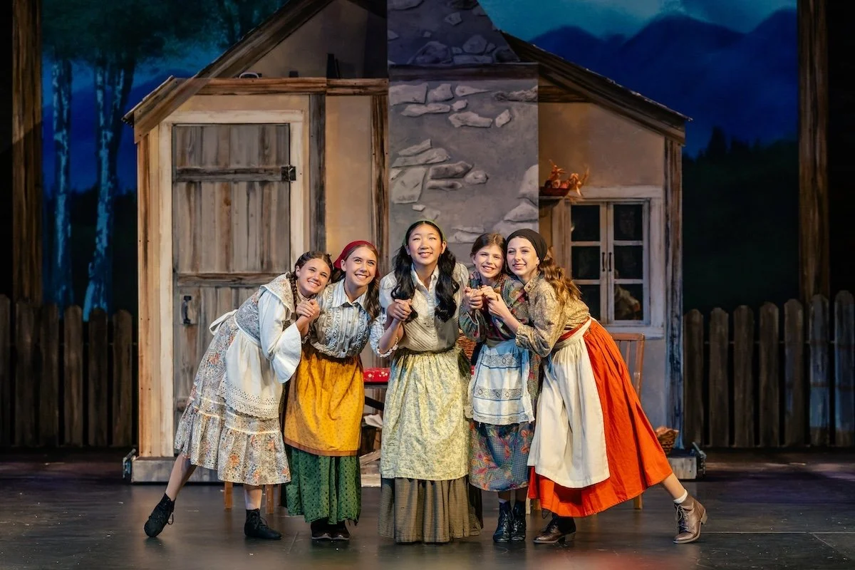 Five young girls in vintage costumes standing on stage in front of a rustic house set, smiling and posing together during a theatrical performance.