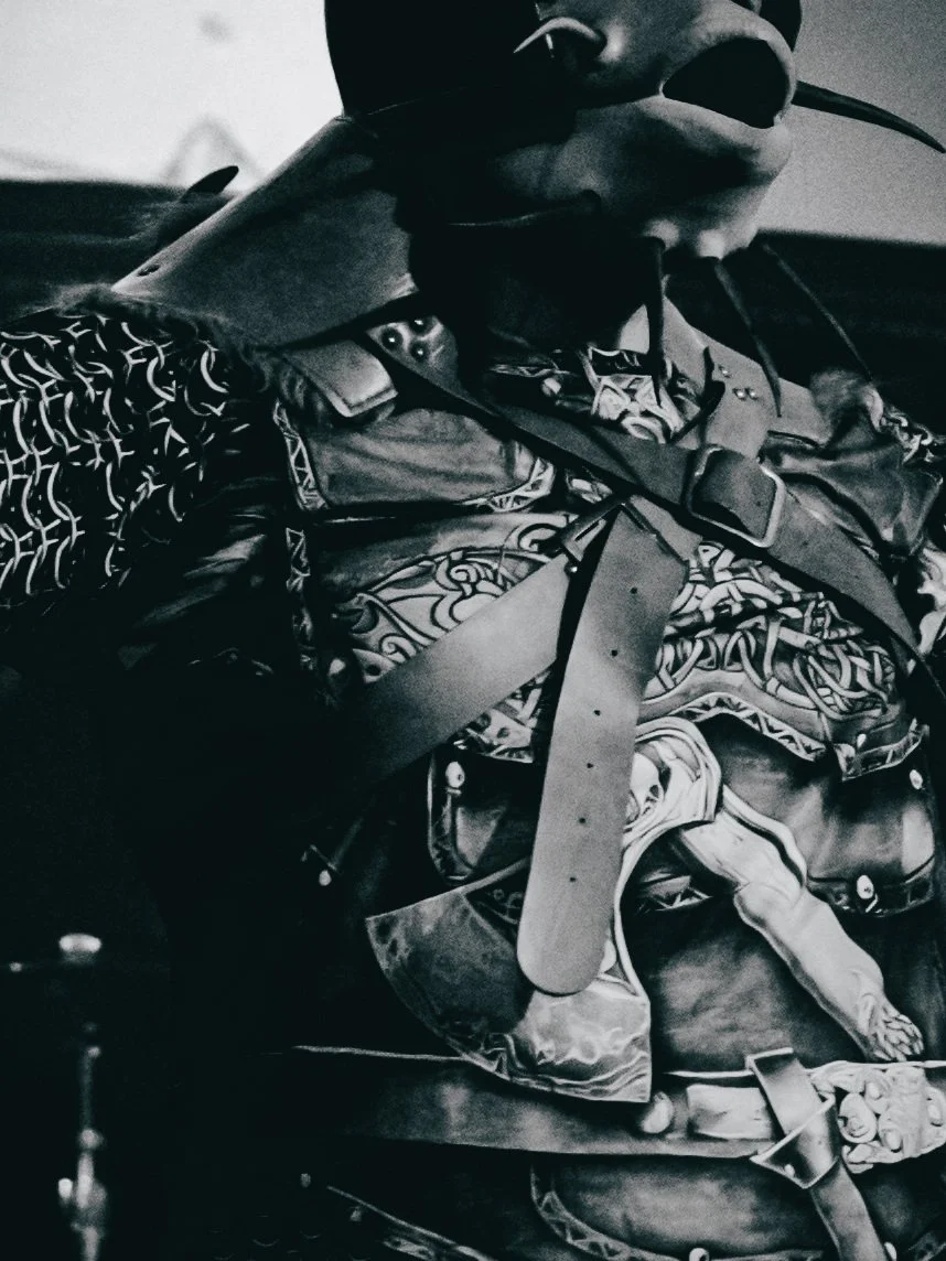 A person wearing sunglasses, a hat, and a patterned shirt with various straps and accessories, lying down or reclining, in a black-and-white photo.