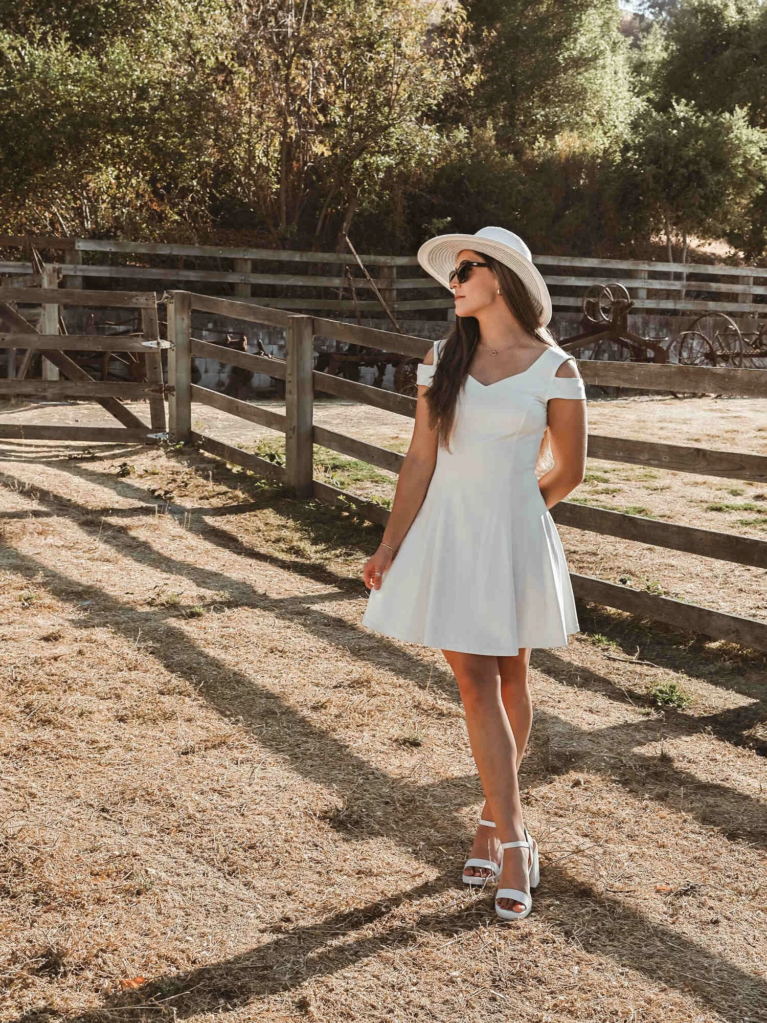 A woman in a white dress, large sun hat, and sunglasses standing outdoors near a wooden fence, with trees and old wagons in the background.