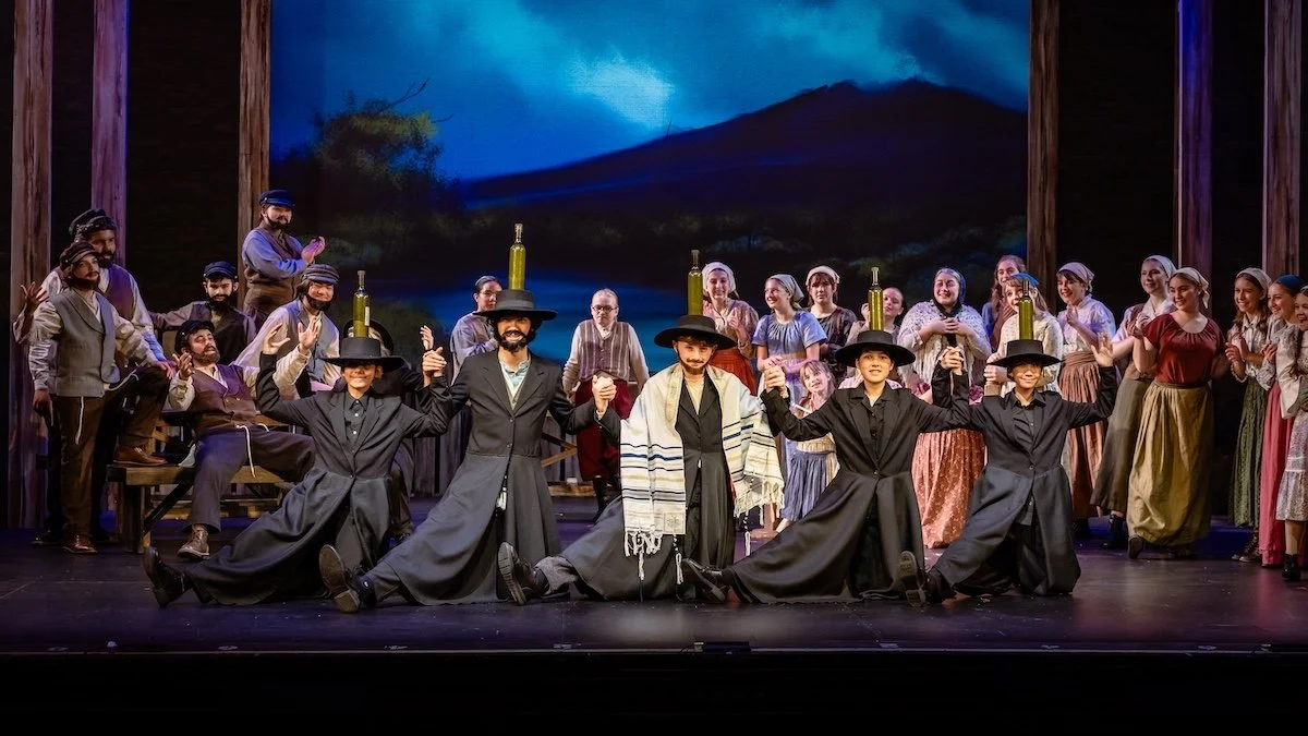 Performers in traditional Spanish costumes on stage, with a mountain backdrop, participating in a dance or celebration.