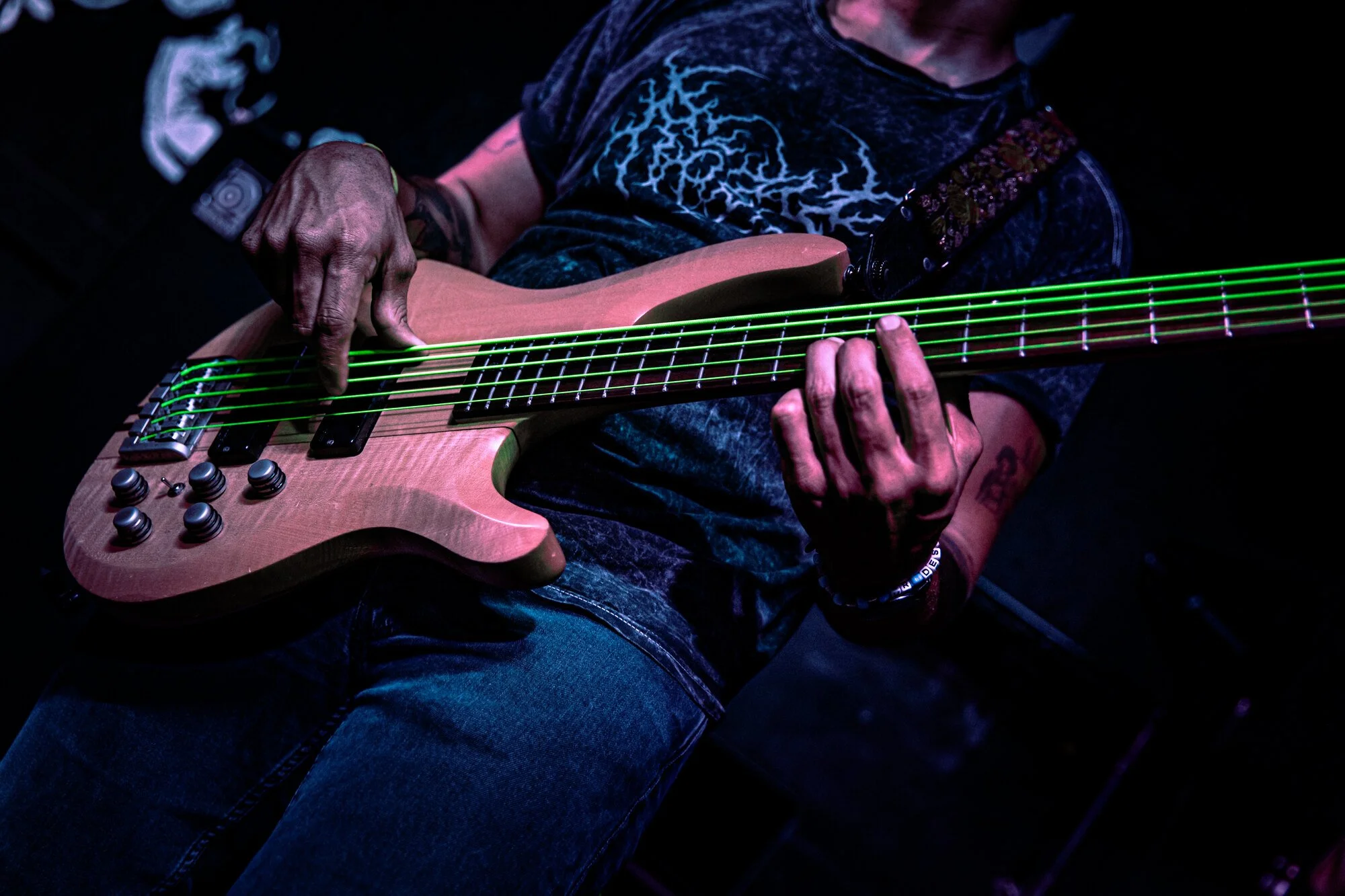 A person playing an electric bass guitar with green laser strings, wearing a dark graphic t-shirt and jeans, in a dark setting.