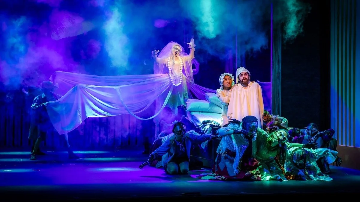 The image shows a theatrical scene with a group of actors on stage. One actor is elevated, wearing makeup and jewelry, dressed as a ghostly or divine figure, holding a flowing fabric. The other actors are kneeling or sitting around a bed, dressed in 
