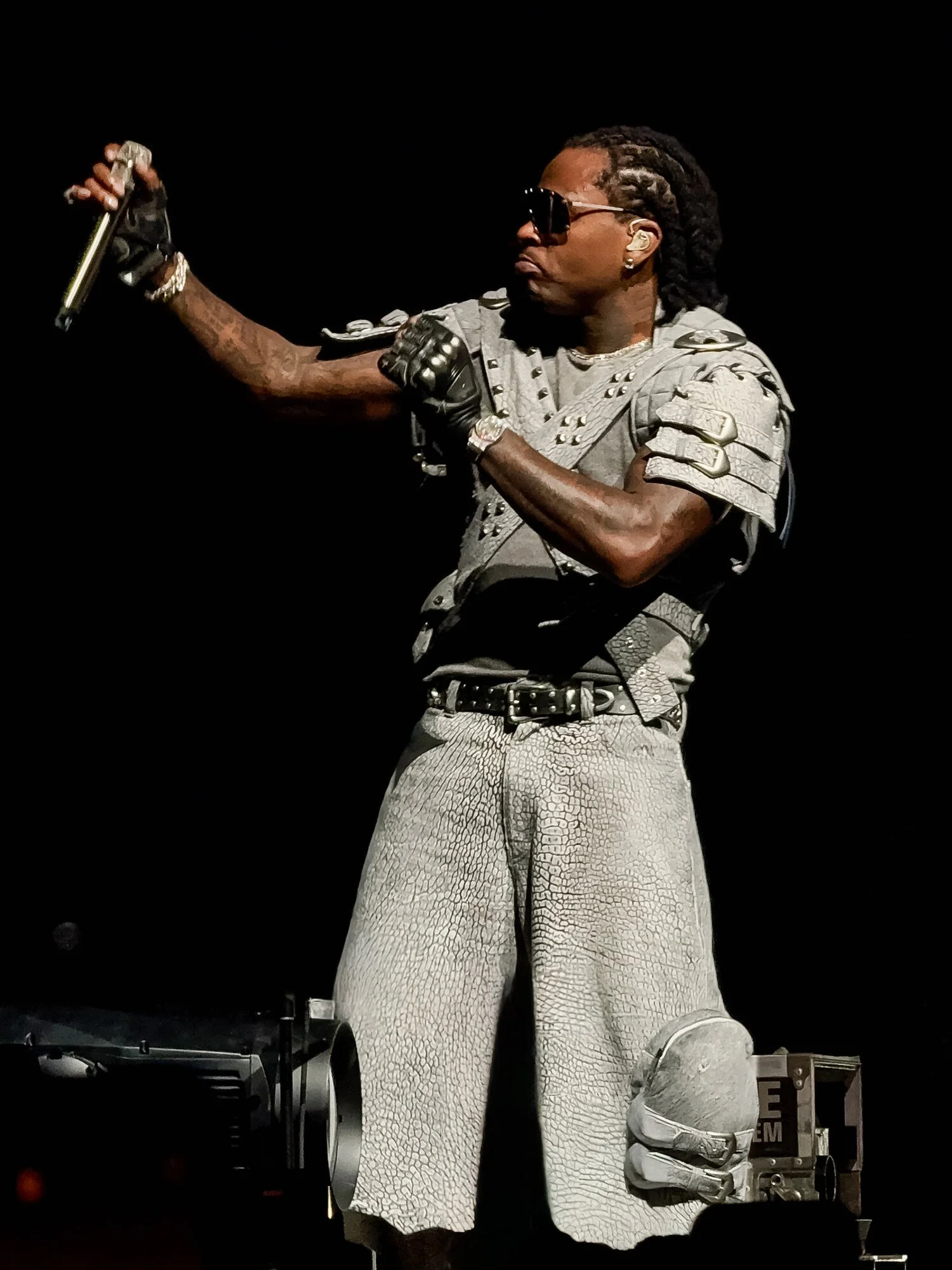 A man wearing sunglasses, a gray armored vest, and patterned pants holding a microphone while performing on a dark stage.