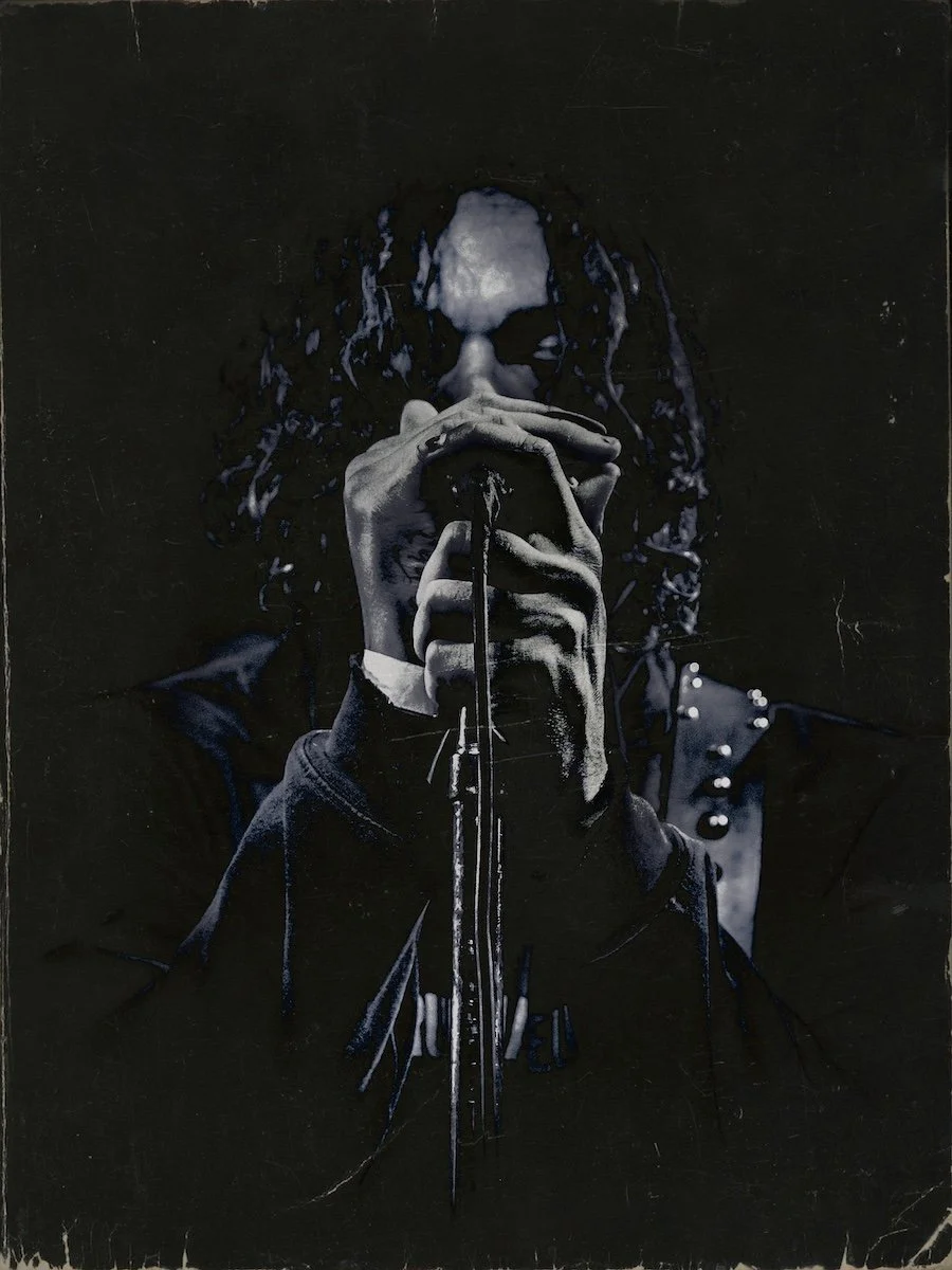 A person with dreadlocks holding a microphone close to their mouth, wearing a dark jacket with metallic studs on the shoulders, against a dark background.