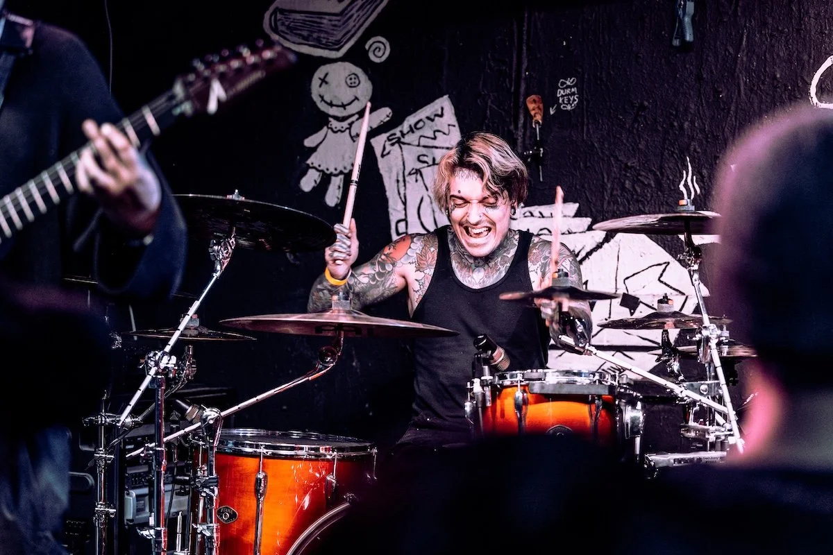 A young man with tattoos on his arms and chest playing drums passionately during a live music performance, with a guitarist partially visible in the foreground and graffiti art on the wall behind him.