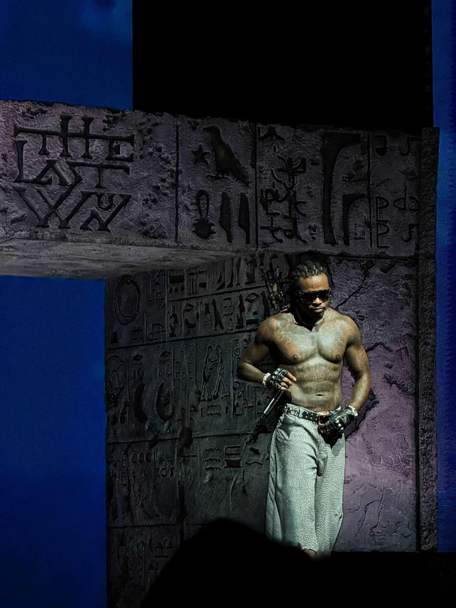 A shirtless man with tattoos, wearing sunglasses, gloves, and light-colored pants, holding a microphone, stands against a wall with Egyptian-style hieroglyphic carvings. Gunna concert.
