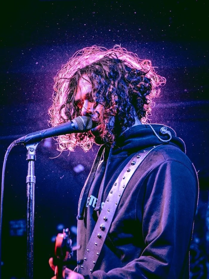 A person with curly hair singing into a microphone on stage, wearing a dark hoodie and a studded guitar strap, with stage lights creating a colorful background.