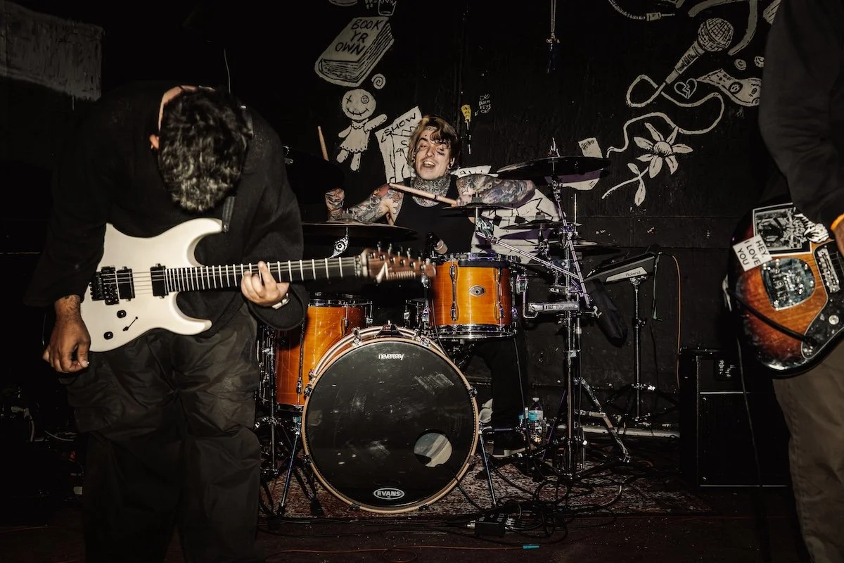 Musicians playing instruments on stage, including a guitarist, drummer, and bassist, with dark background and artwork on the wall.
