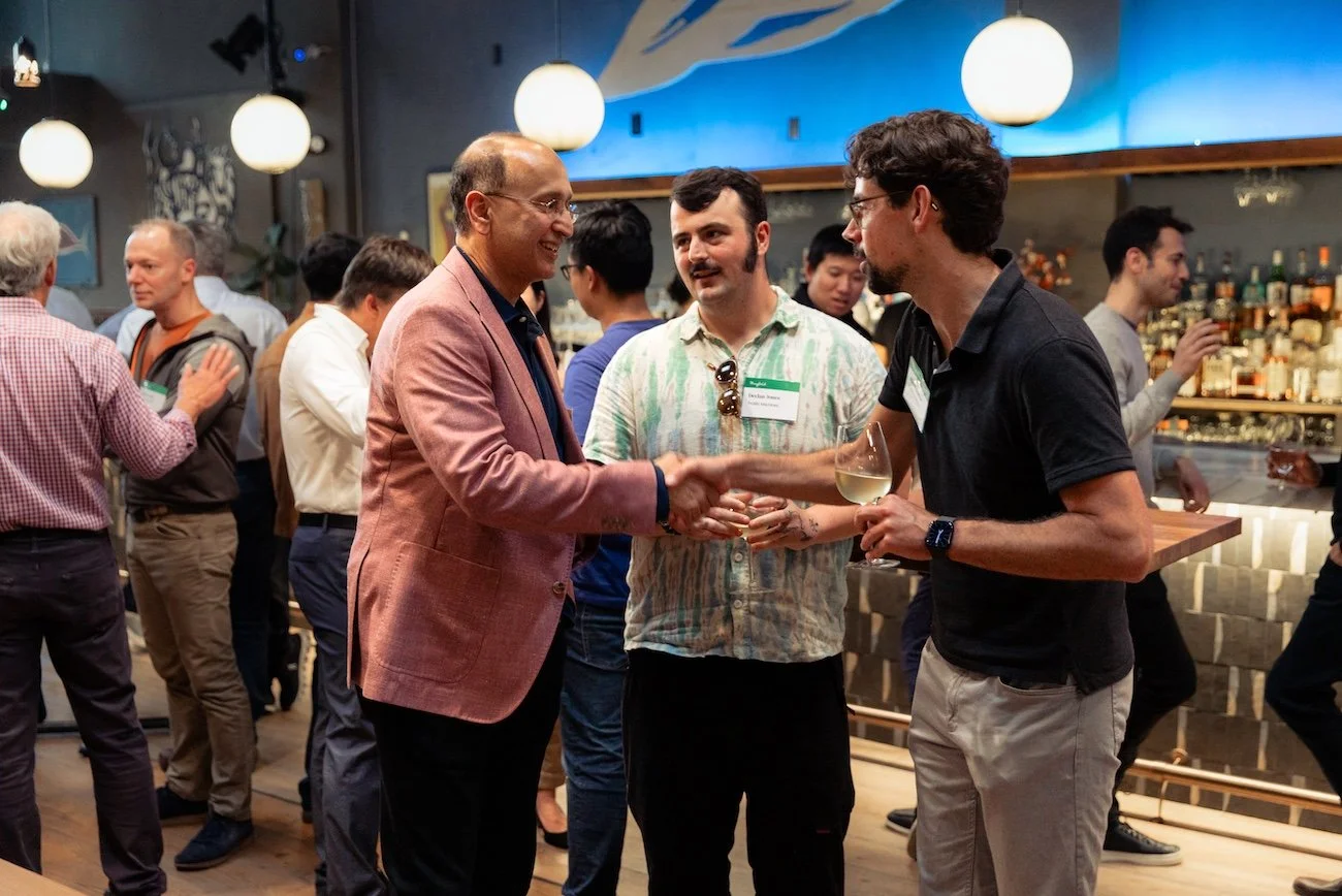 People at a networking event in a bar, engaging in conversations. A man in a pink blazer shakes hands with a man in a black polo holding a glass of white wine.