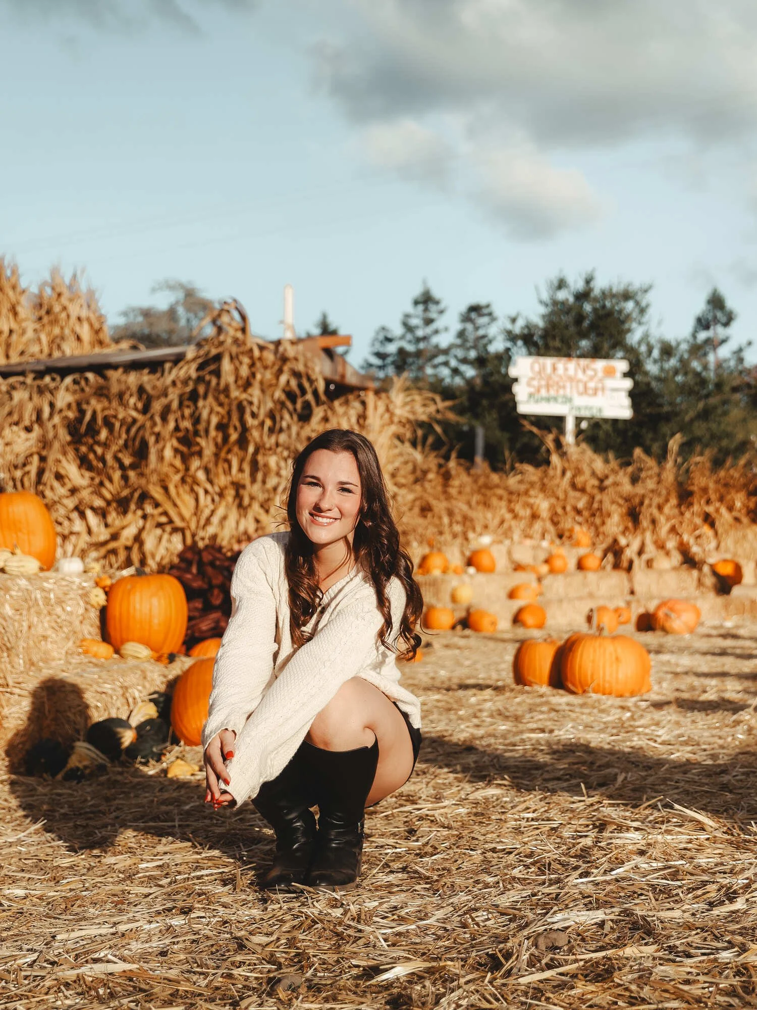 A young woman crouching outdoors among pumpkins at a pumpkin patch during fall.
