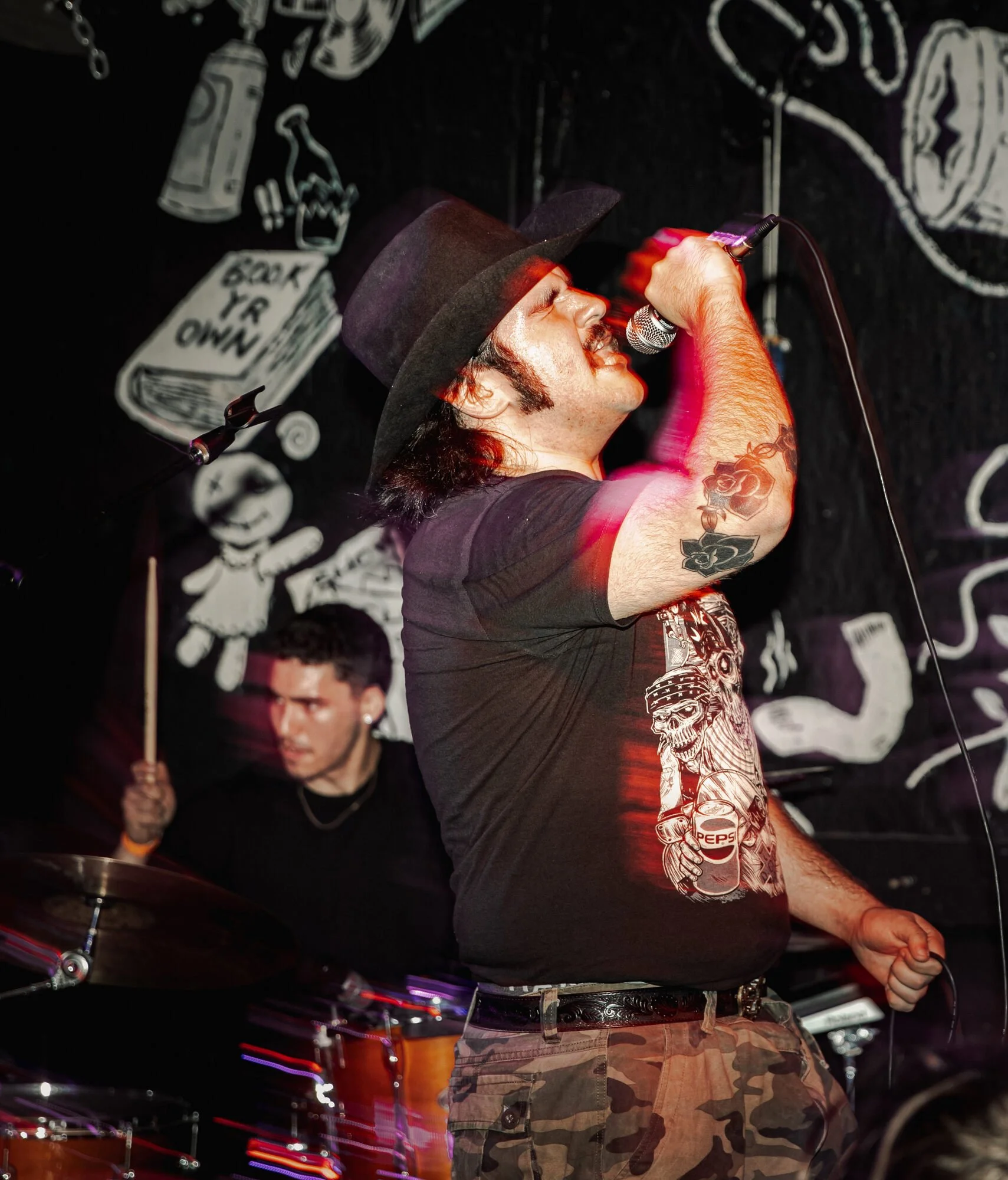 A male singer in a black t-shirt, camouflage pants, a wide-brimmed hat, and tattoos on his arm, performing with a microphone on stage. A drummer with dark hair and earrings plays in the background, with a black wall with white graffiti and drawings.