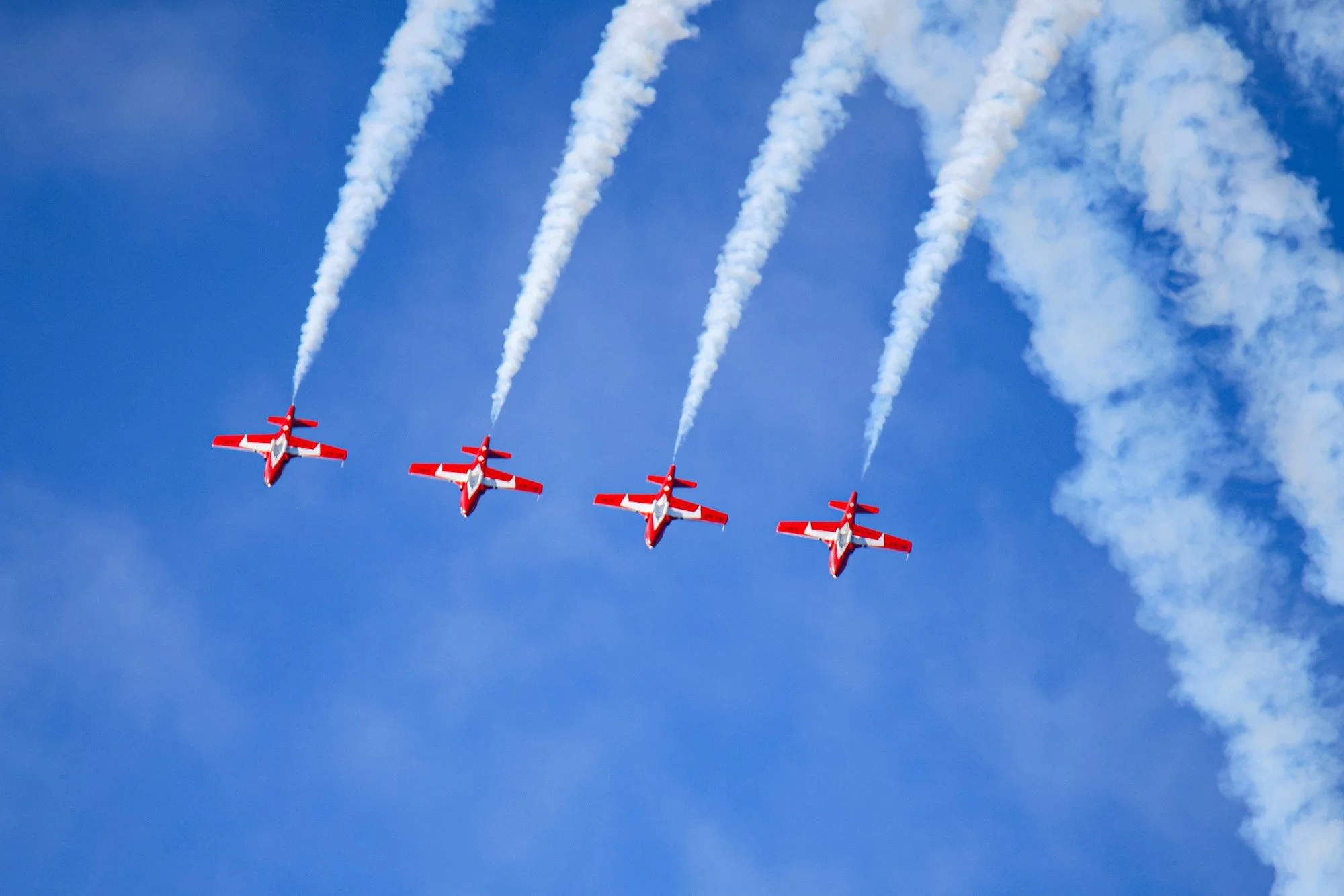 Four red and white airplanes flying in formation with white smoke trails in a blue sky.