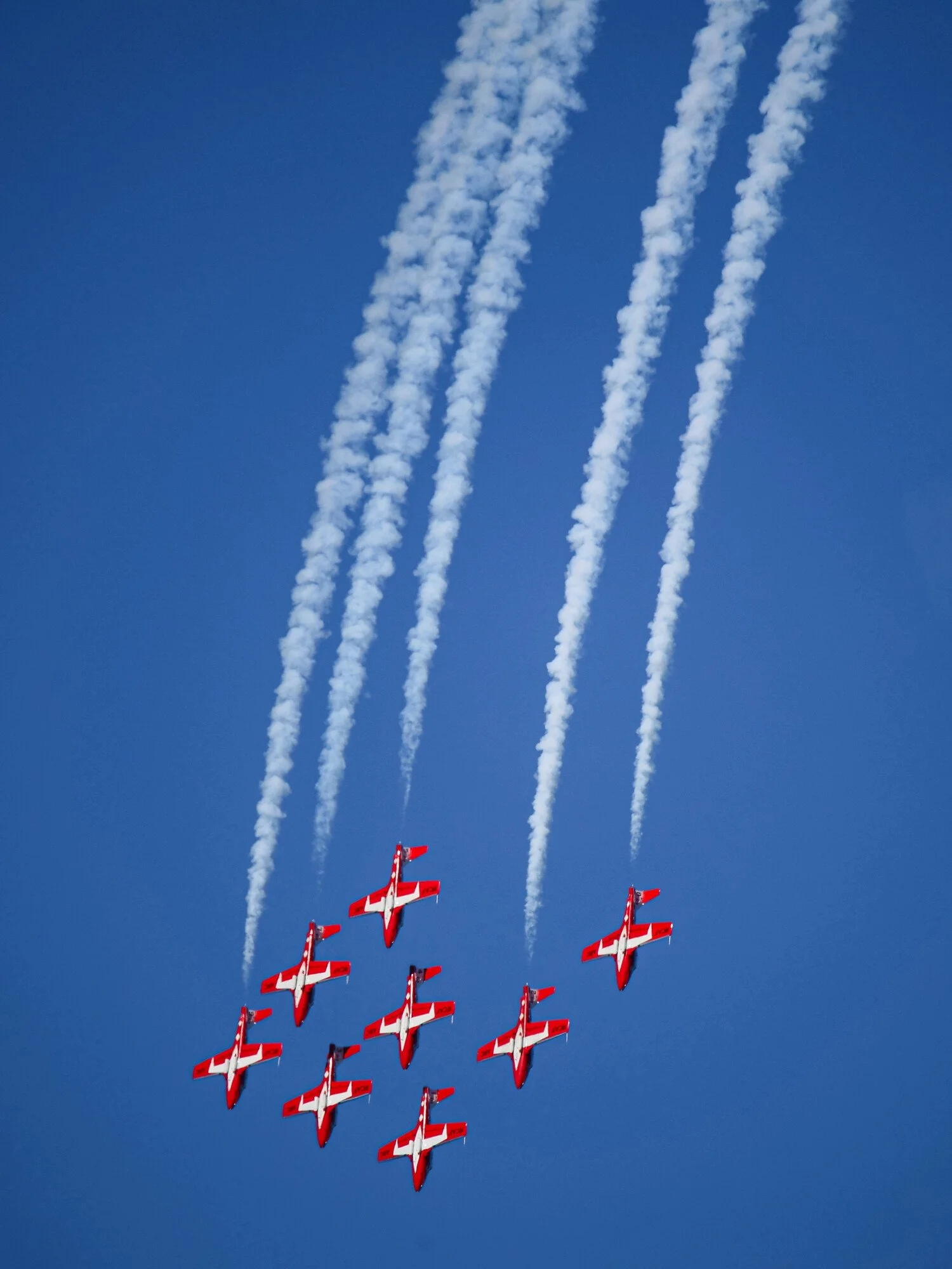 A formation of ten red and white airplanes flying in a delta pattern against a blue sky, leaving white smoke trails behind.