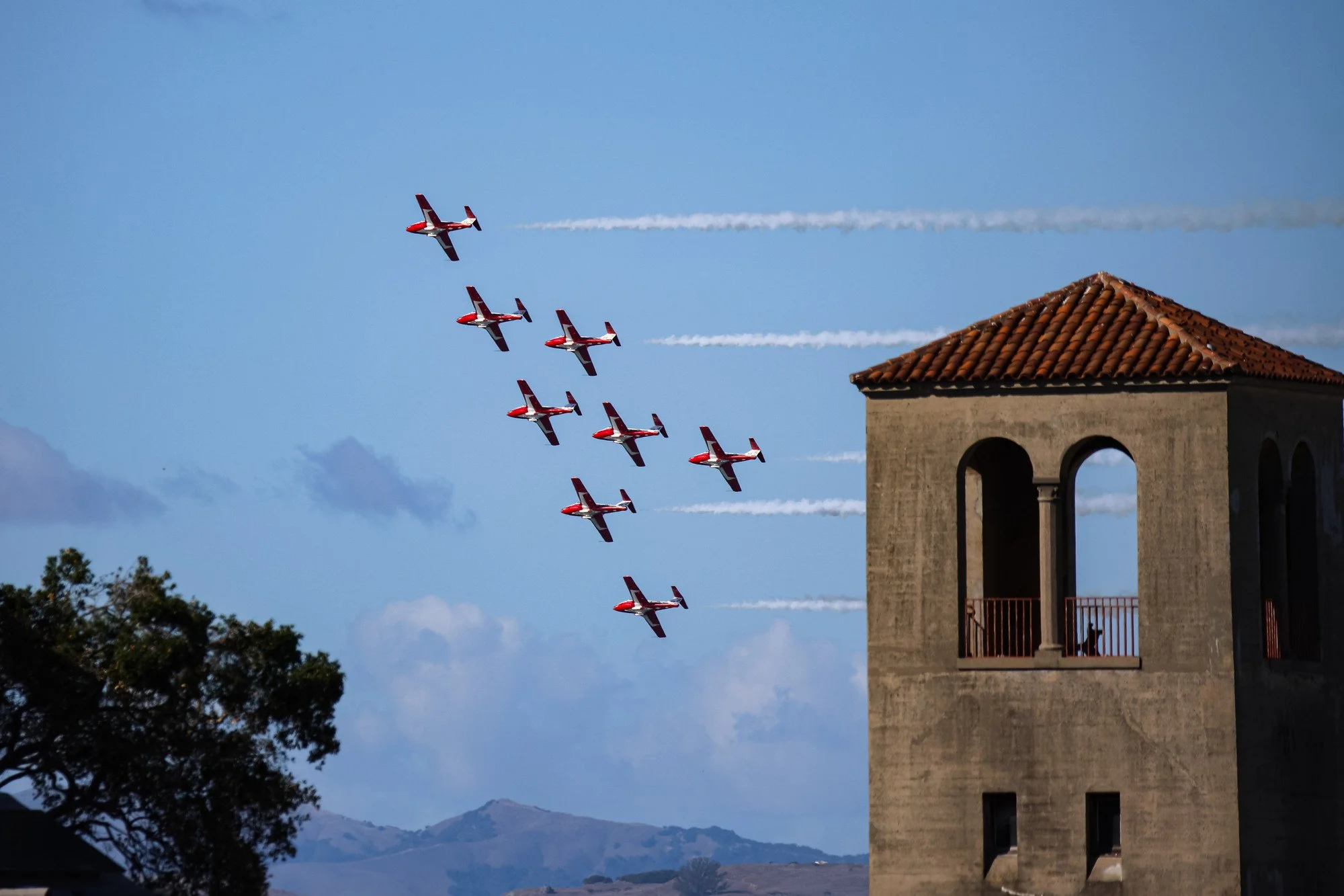 A formation of red and white aircraft flying in the sky, leaving white smoke trails behind them, near a stone building with a red-tiled roof and bell tower, with mountains in the background.