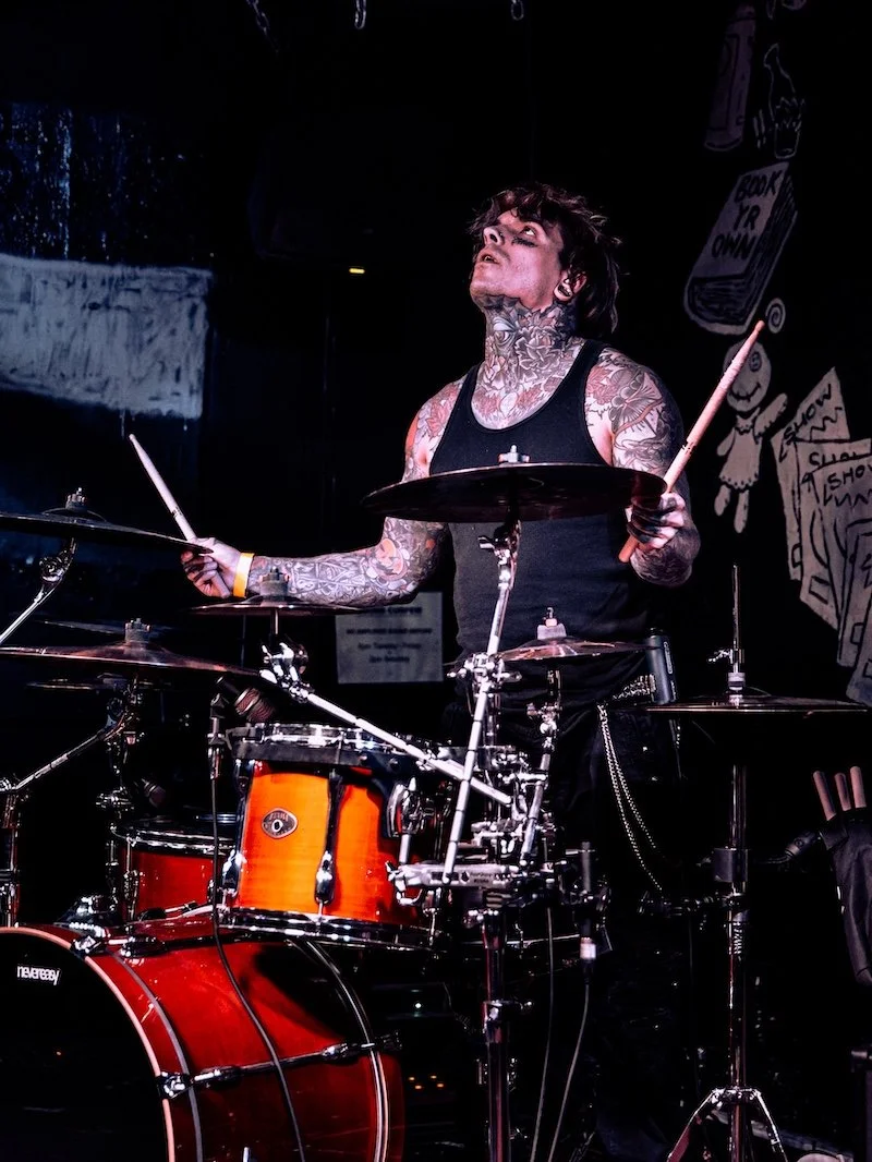 A tattooed man playing drums on stage, wearing a black tank top, with a dark background and sketches on the wall.