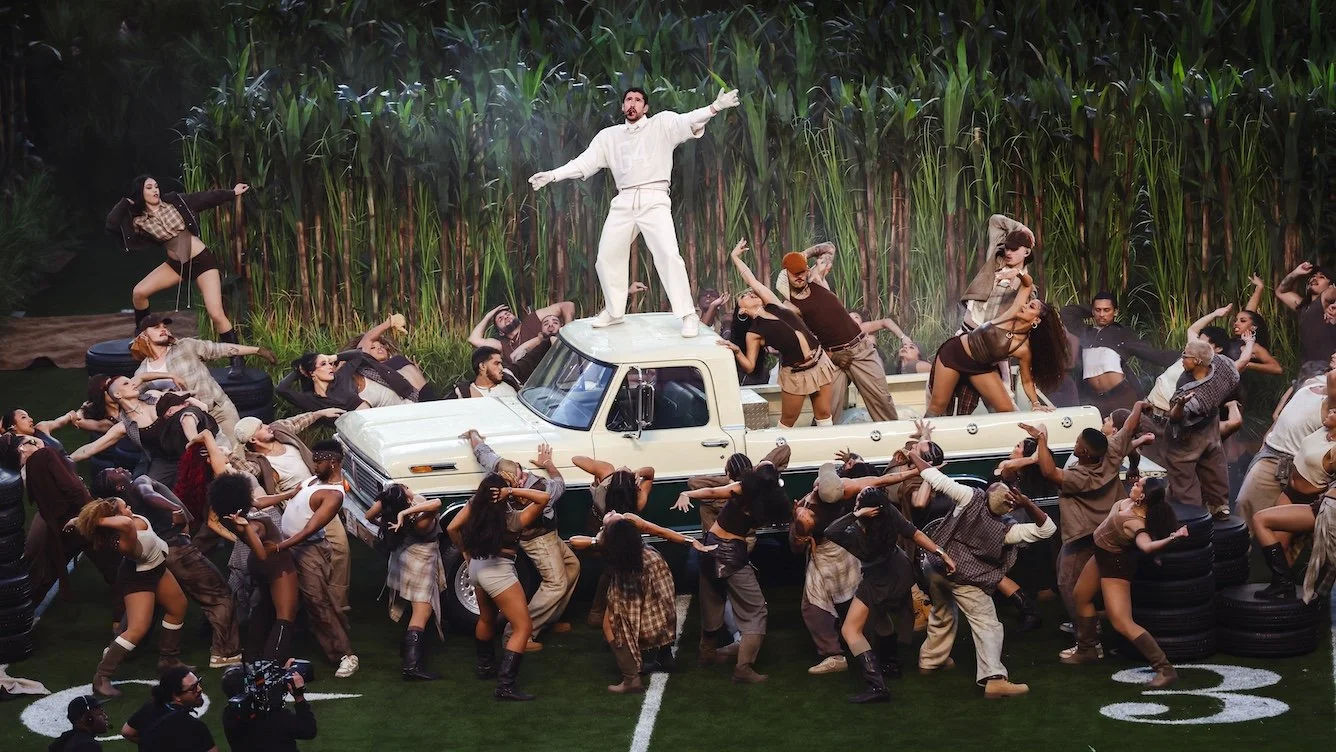 Bad Bunny at the Super Bowl LX Halftime show, in an all-white outfit stands on top of a vintage pickup truck, singing during a high-energy halftime show as dozens of dancers surround the truck on a field-themed stage with tall green reeds in the back
